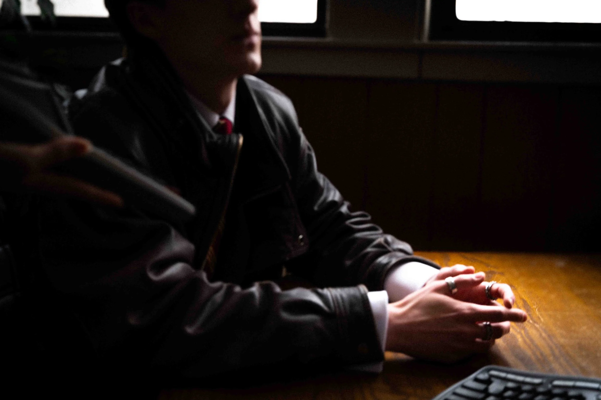 A person in a dark leather jacket sitting at a wooden table with their hands clasped, illuminated by light near a window, with a computer keyboard partially visible in the foreground.