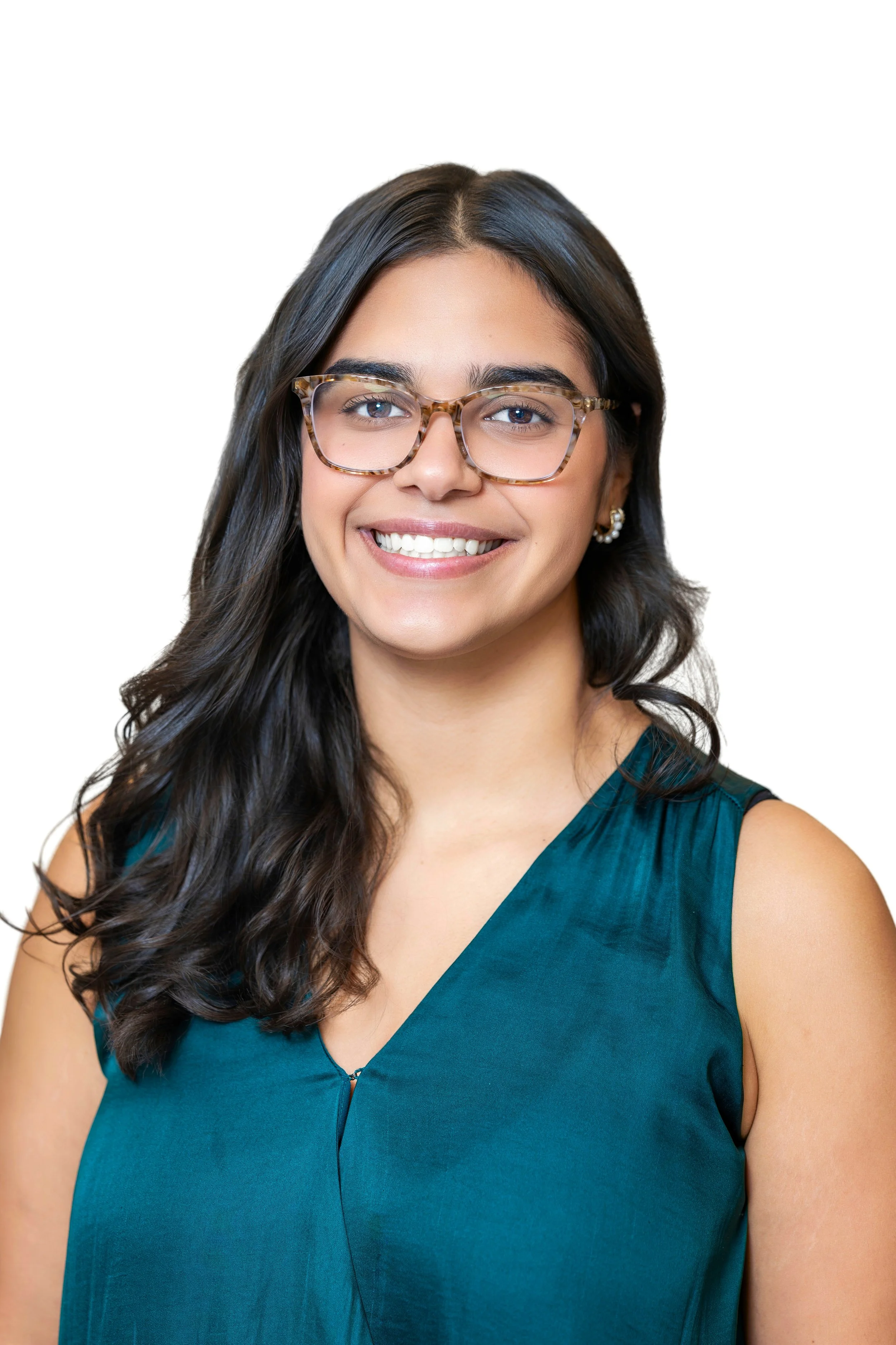 A woman with dark hair, glasses, and jewelry smiling in front of a light-colored wall.