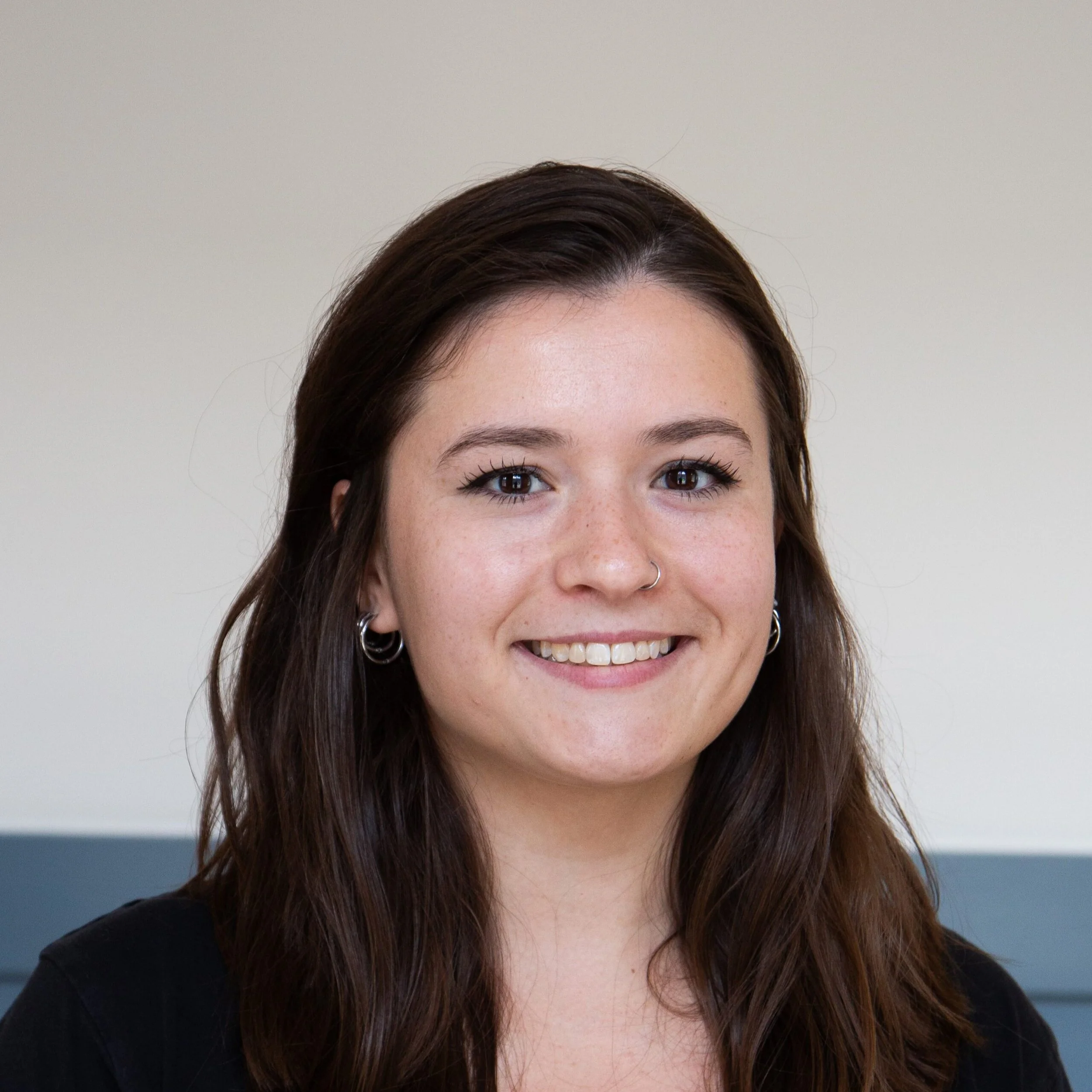 Close-up of a woman's face with brown hair, smiling, wearing a silver nose ring and earrings, against a neutral background.