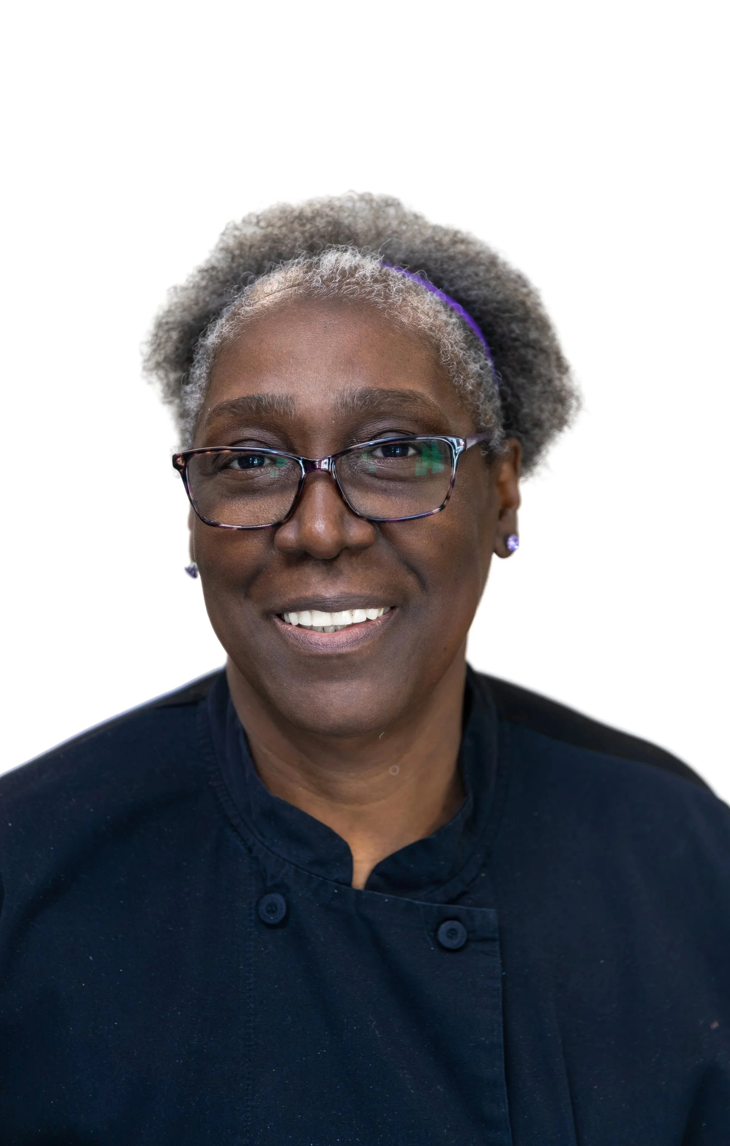 Close-up of a smiling woman with short gray hair, glasses, and earrings, wearing a dark shirt, standing against a pastel blue and white background.