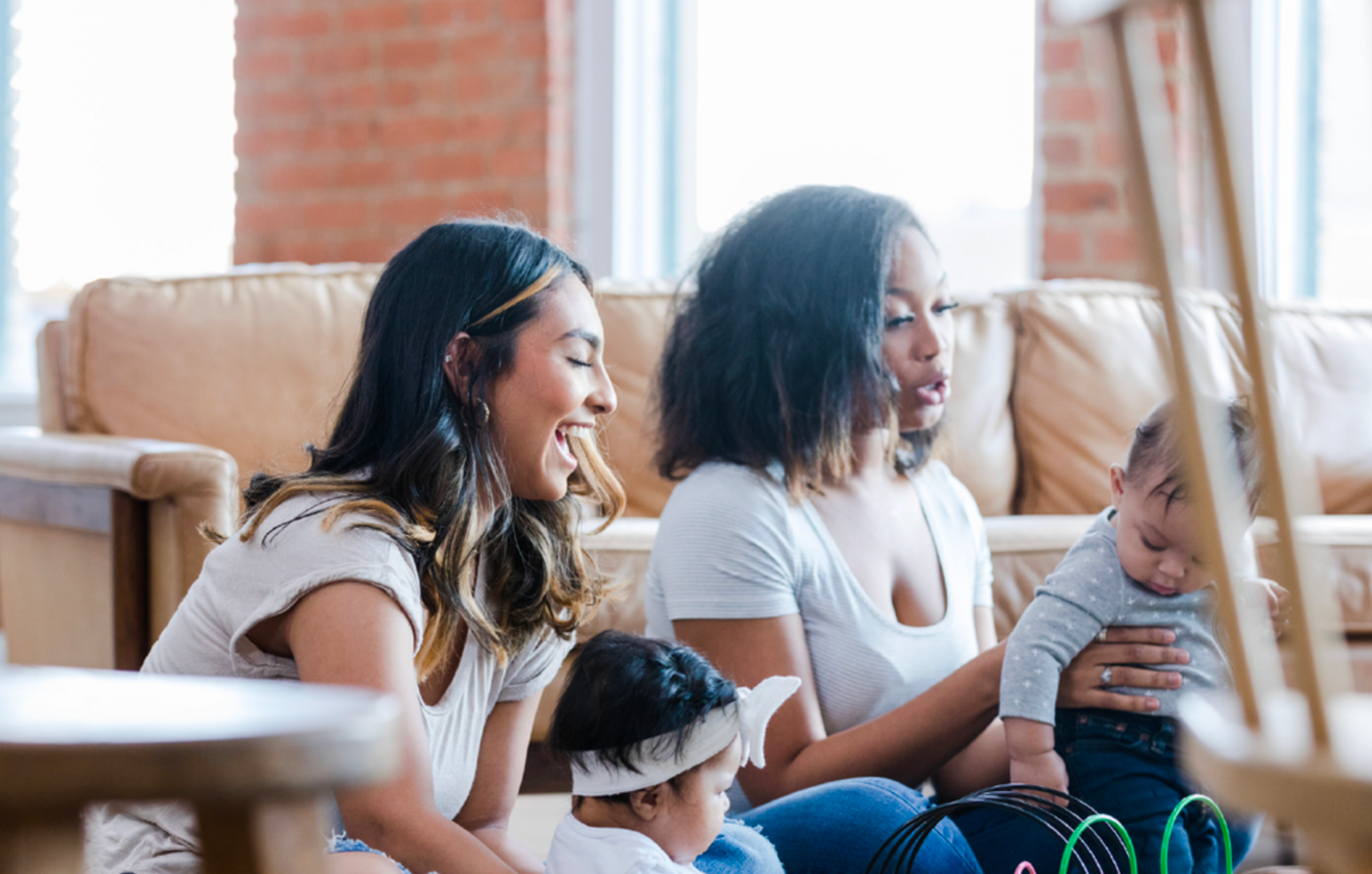 Two women and two children sitting on the floor in a bright room with large windows and exposed brick walls, engaging with each other.