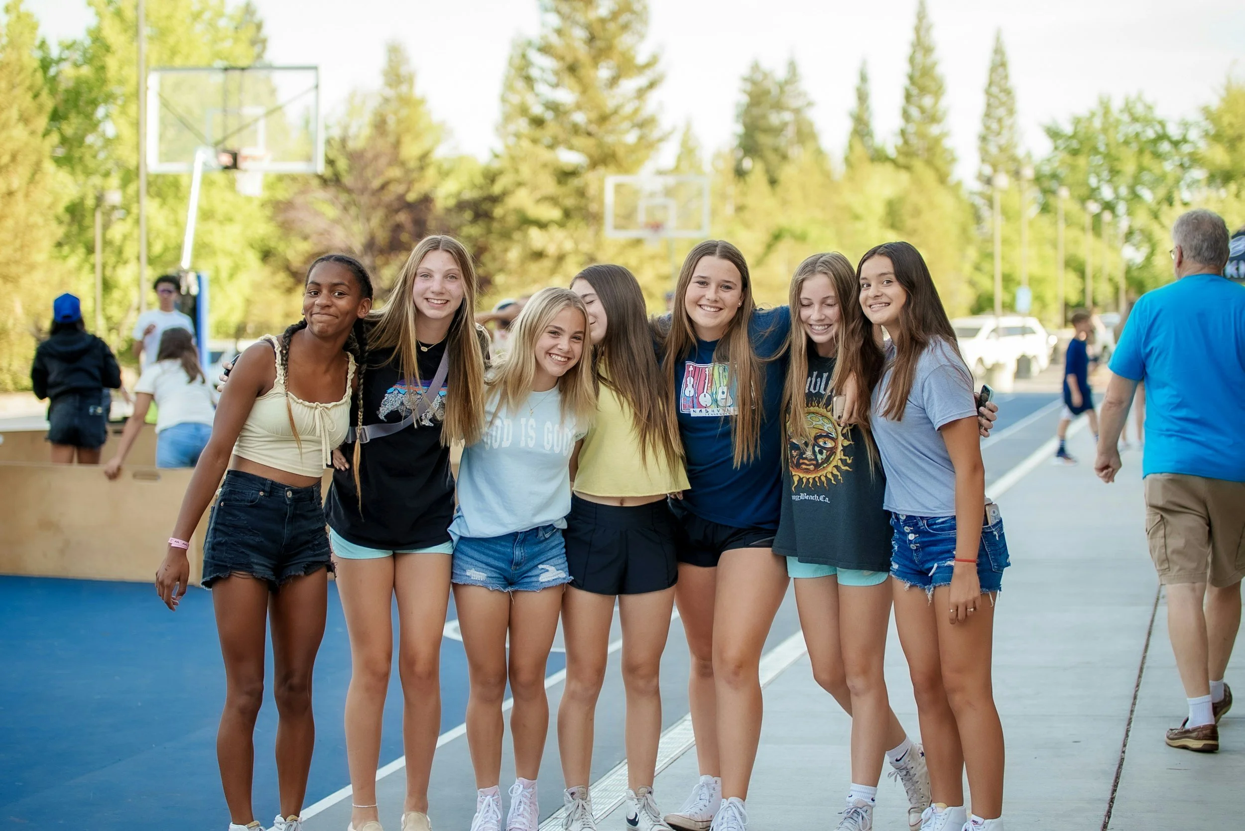 A group of eight teenage girls smiling and walking together on a basketball court outdoors, with trees and a basketball hoop in the background.