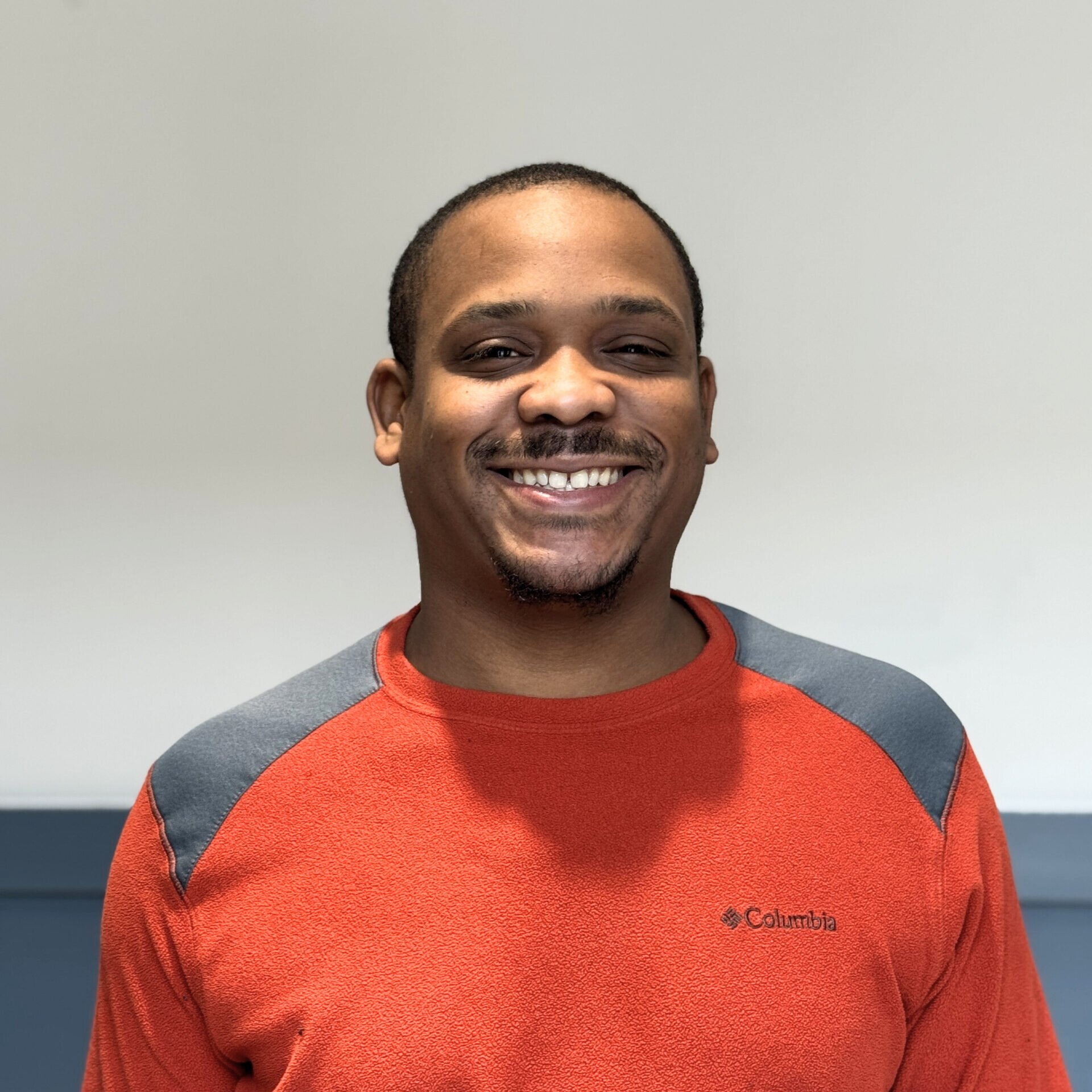 A smiling man with short hair and a goatee, wearing a red Columbia sweatshirt with gray and black accents, standing against a plain wall.