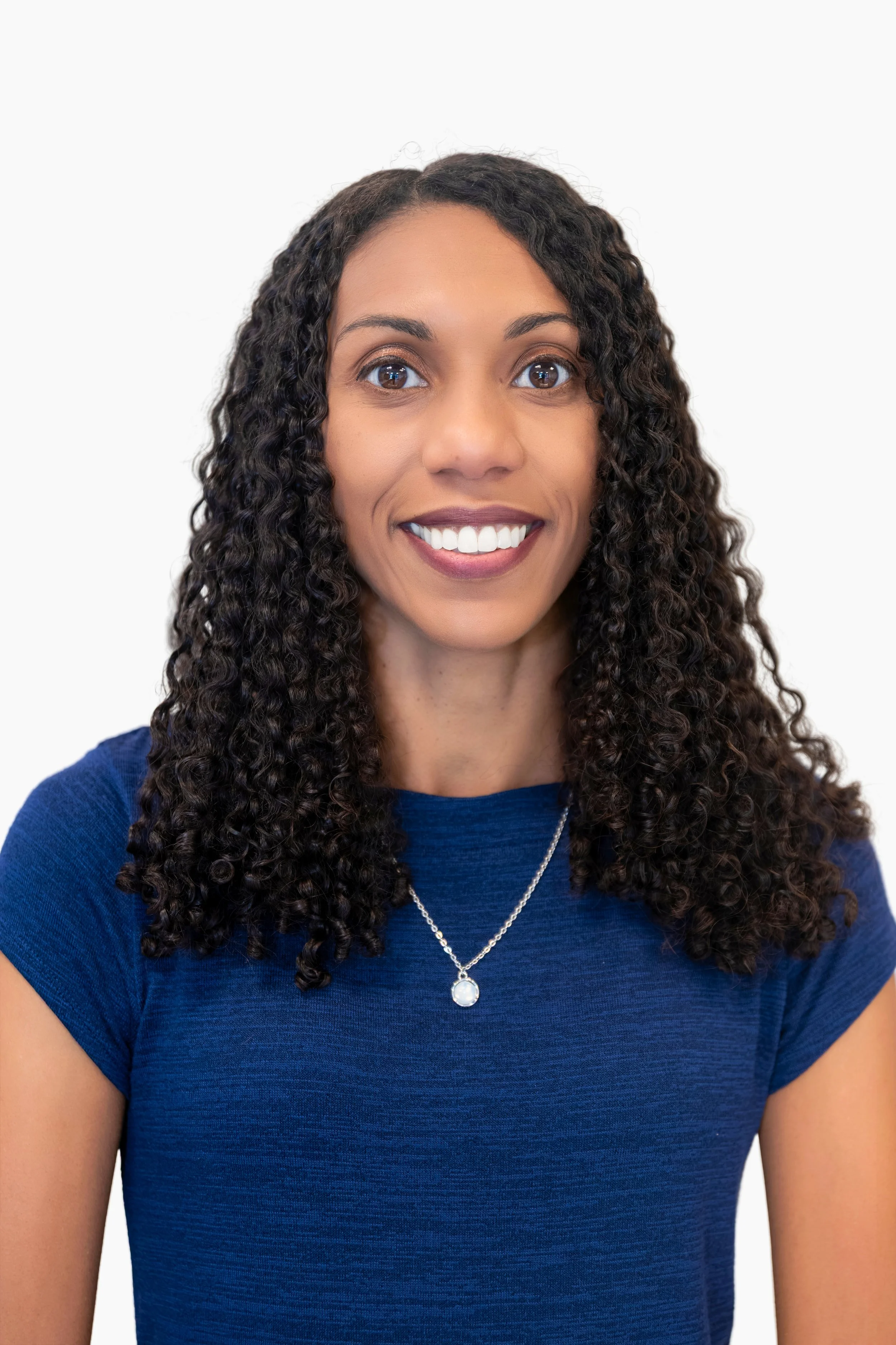 A woman with curly black hair smiling, wearing a black blazer and maroon top, against a plain white background.