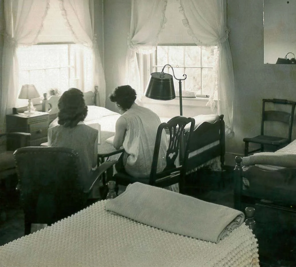 Two women sitting on a bed in a bedroom with vintage furniture, white curtains, and a lamp.