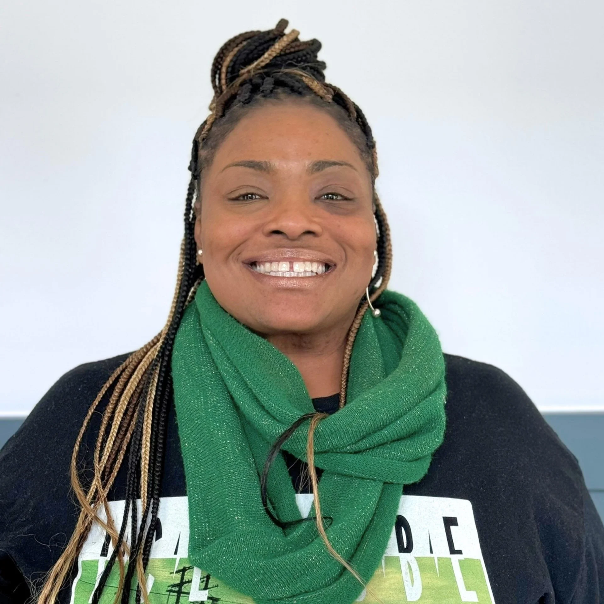 Close-up of a smiling African American woman with braided hair styled in an updo, wearing a green sweater and earrings, against a plain light-colored background.