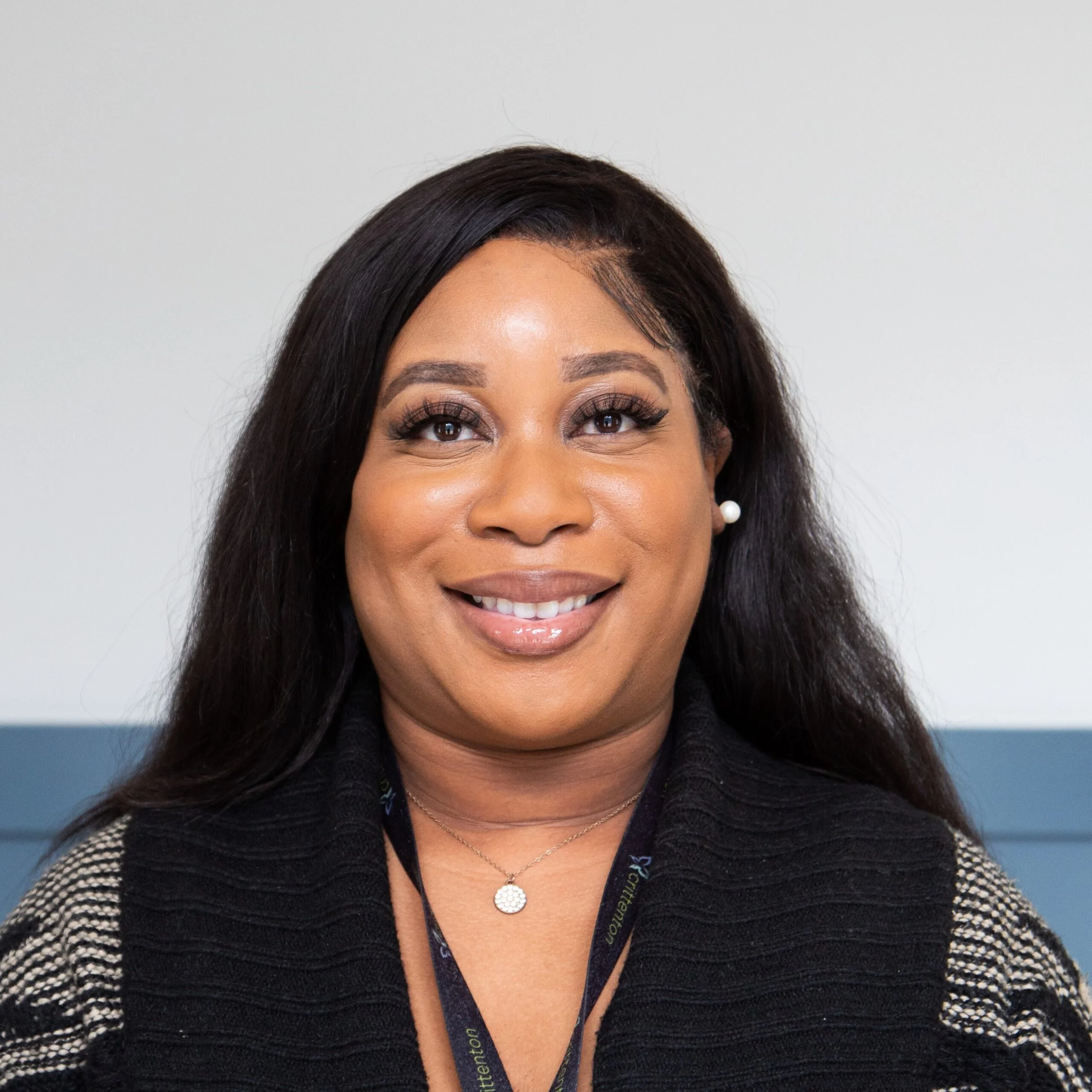 Close-up of a woman with dark hair, smiling, wearing pearl earrings, a layered necklace, and a black and white patterned top, against a light background.