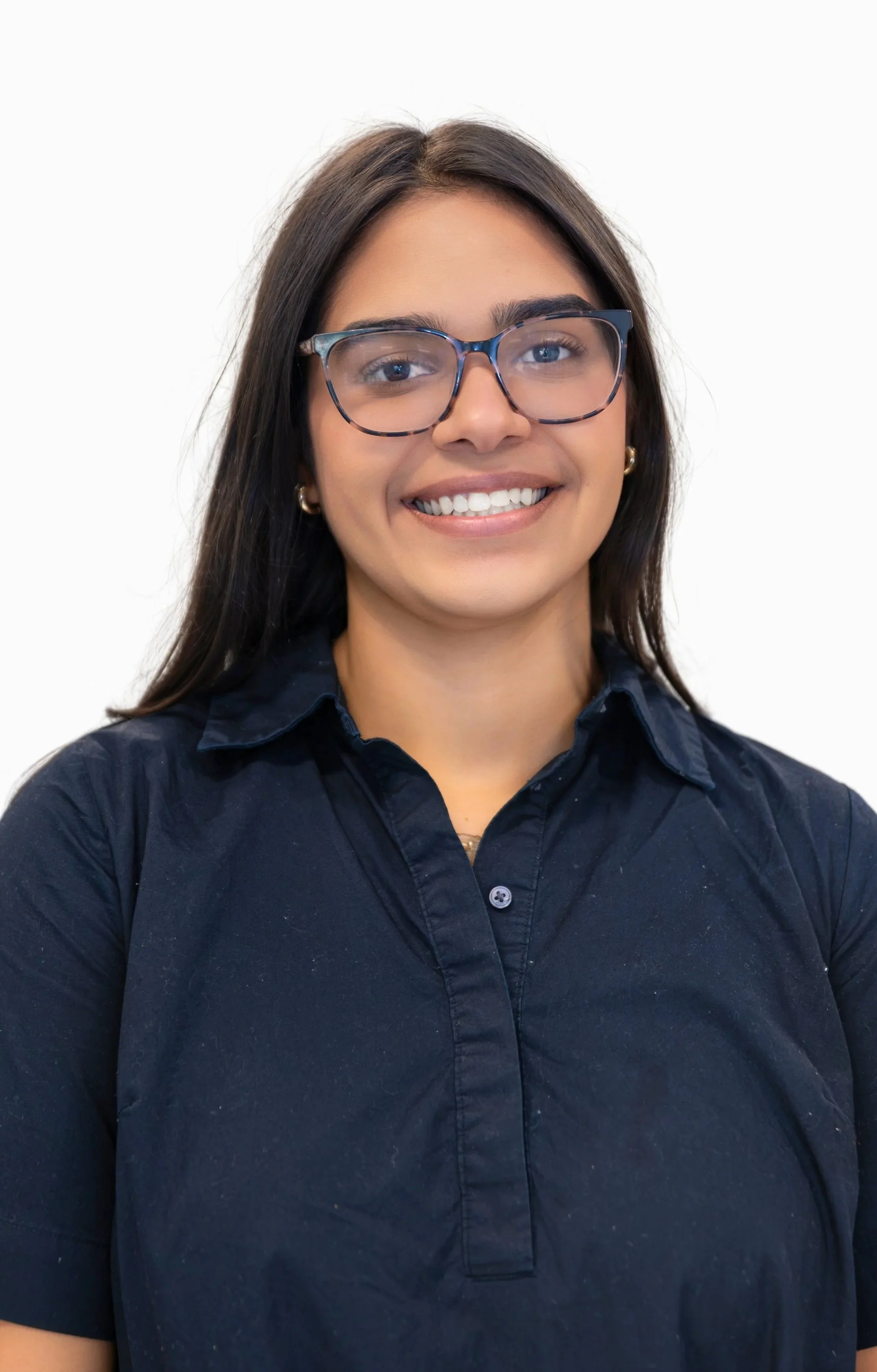 A woman with dark hair, glasses, and jewelry smiling in front of a light-colored wall.