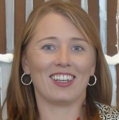 Close-up of a smiling woman with reddish hair and hoop earrings, standing against a wooden background.