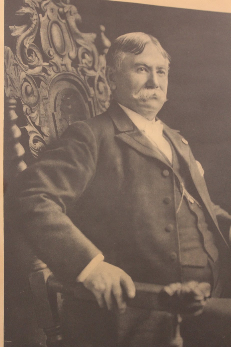 A black-and-white photograph of a man with a mustache, dressed in a suit, sitting in an ornate wooden chair.