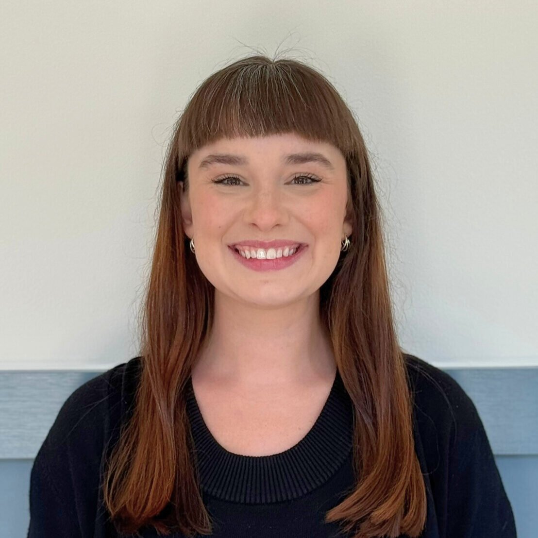 Portrait of a smiling woman with long red hair, wearing a black top and earrings, standing against a light-colored wall.
