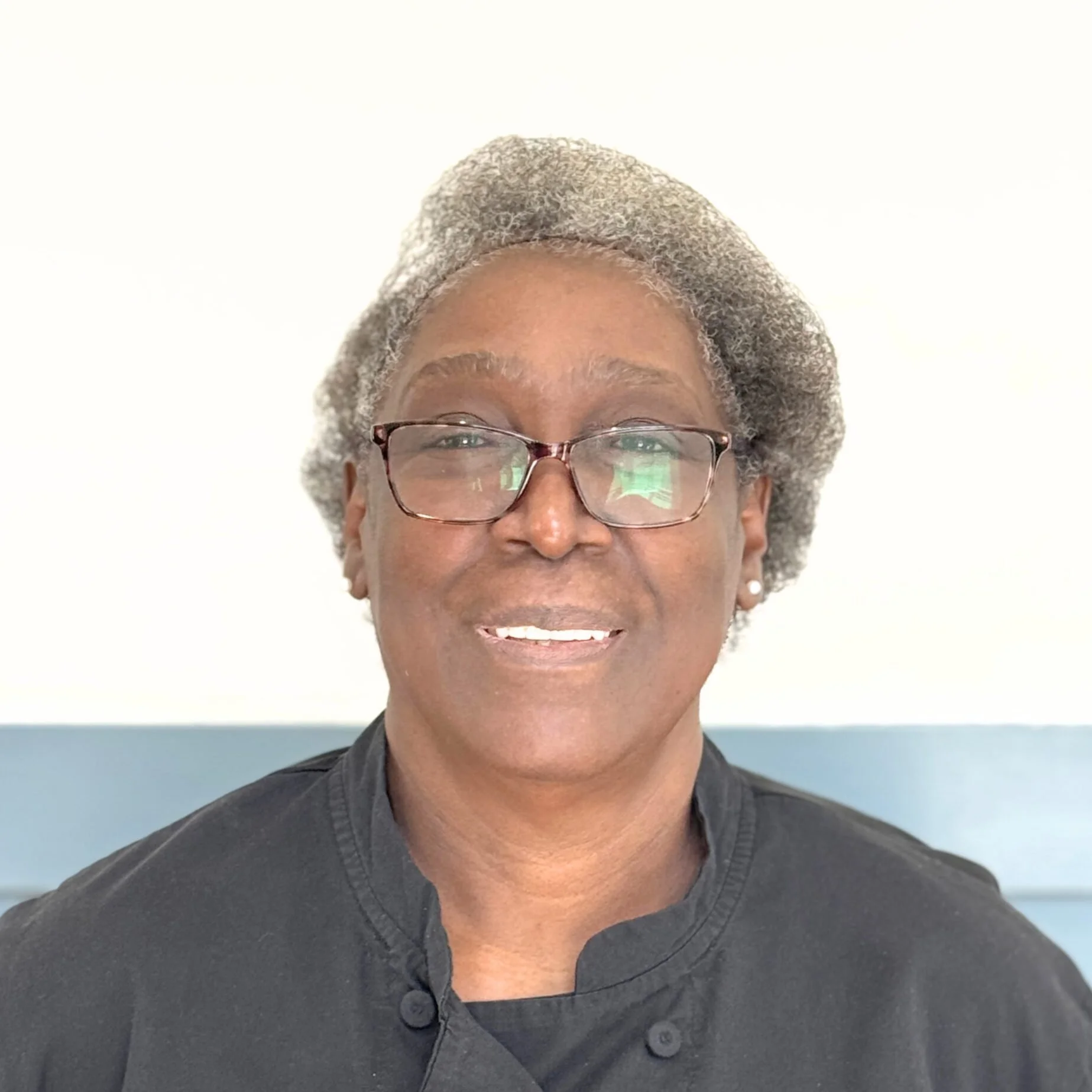 Close-up of a smiling woman with short gray hair, glasses, and earrings, wearing a dark shirt, standing against a pastel blue and white background.