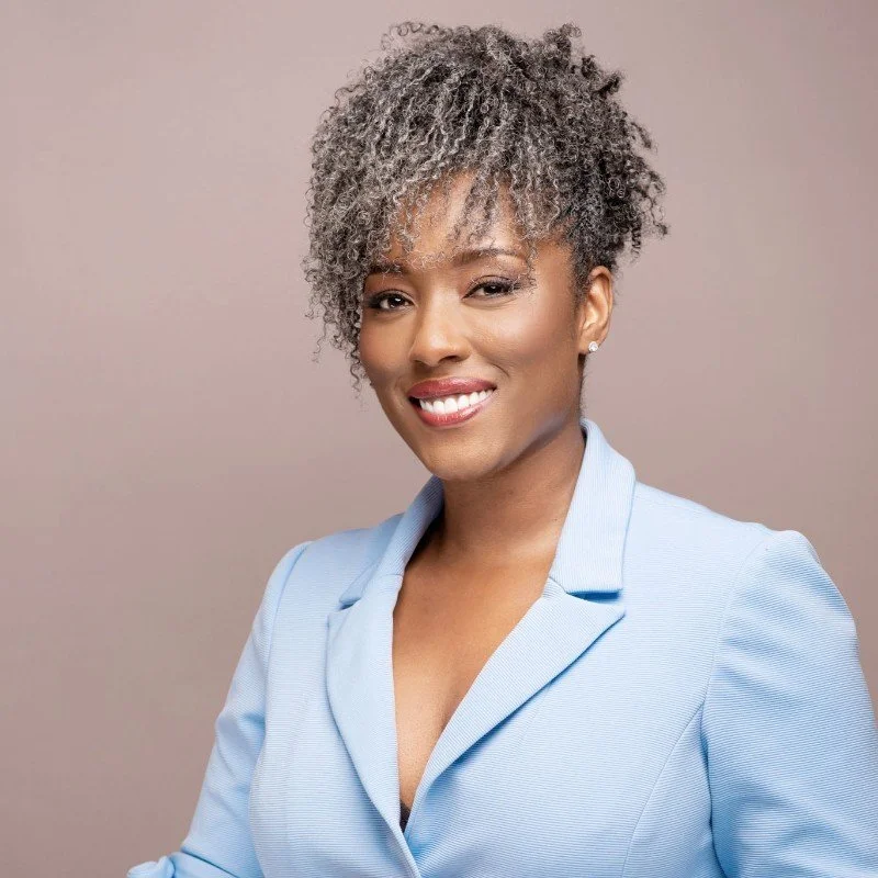 A woman with short, curly, gray and black hair, smiling and winking, wearing a light blue blazer and pearl earrings, against a plain beige background.