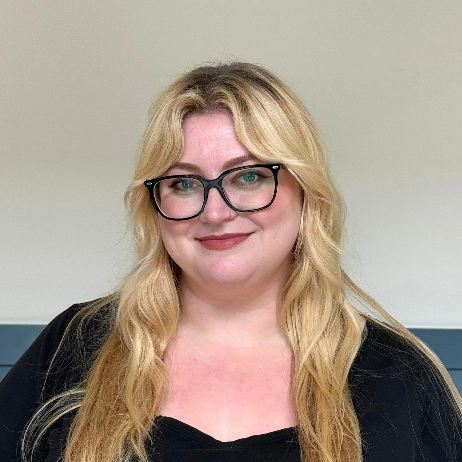 A woman with long blonde hair, glasses, and a black top smiling against a plain light-colored background.