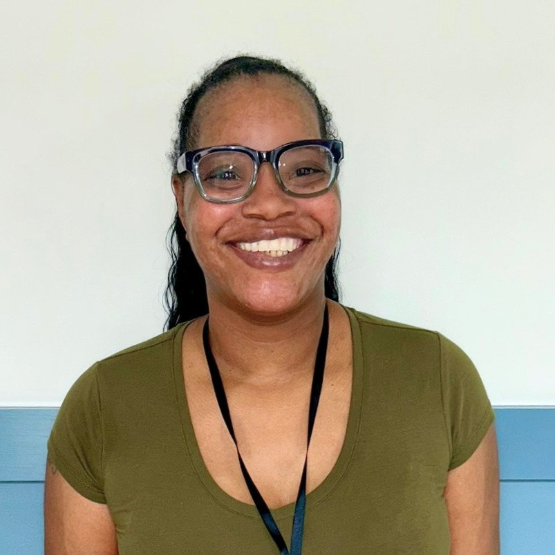 Smiling woman wearing glasses, green T-shirt, and a black lanyard in front of a white and blue wall.