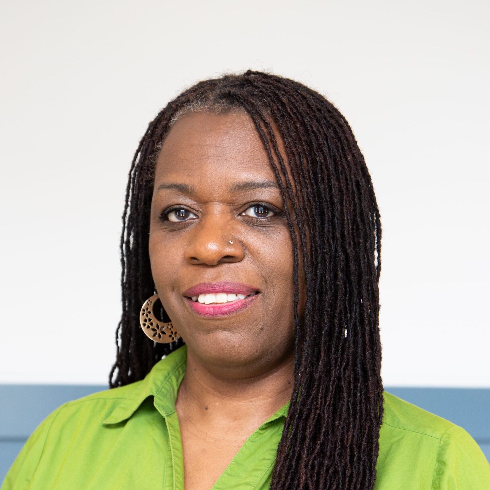 Close-up of an African American woman with dreadlocks, wearing a bright green shirt, and large earrings, smiling against a plain background.