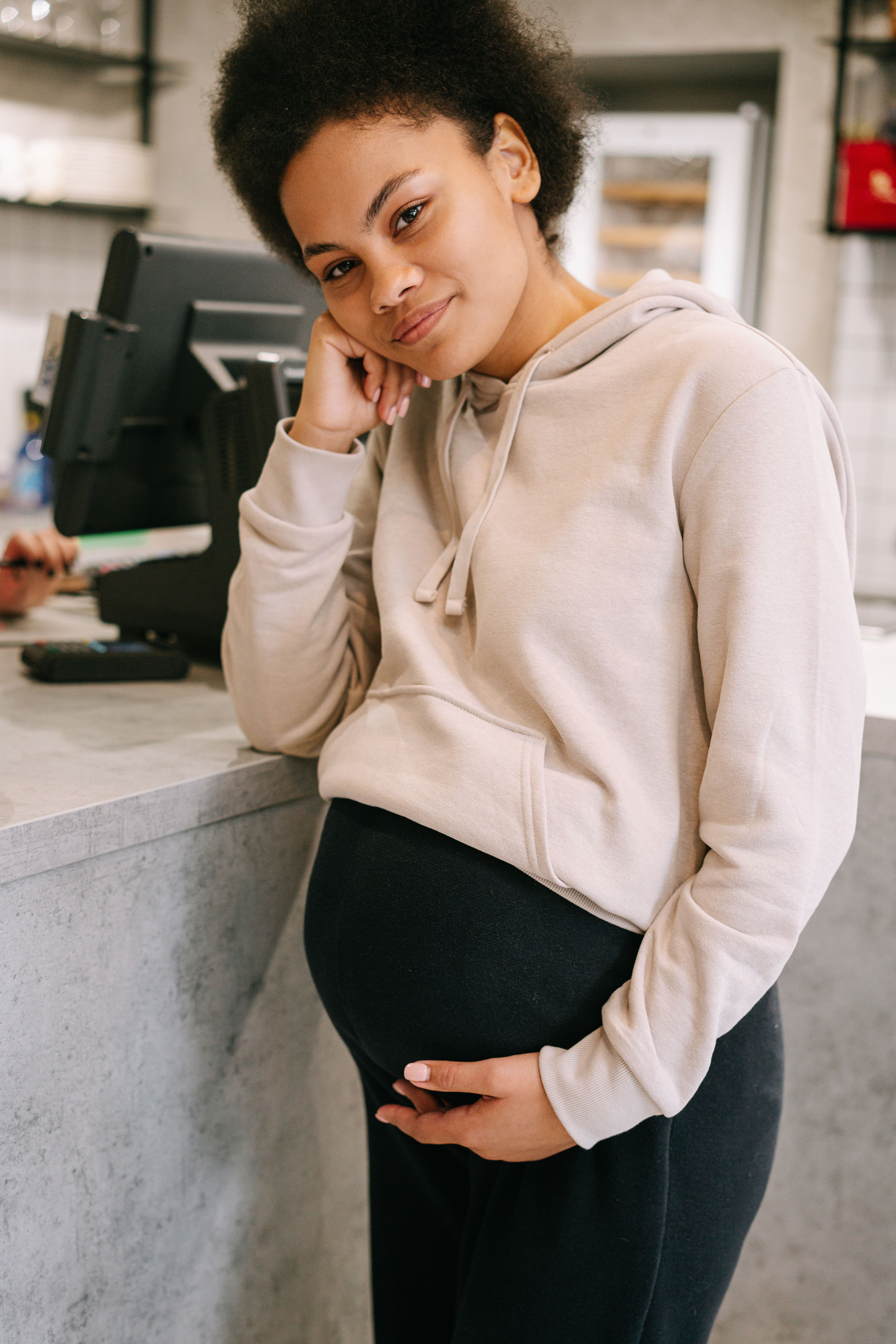 A pregnant woman standing at a counter, gently cradling her belly with her right hand, wearing a beige hoodie and black leggings.