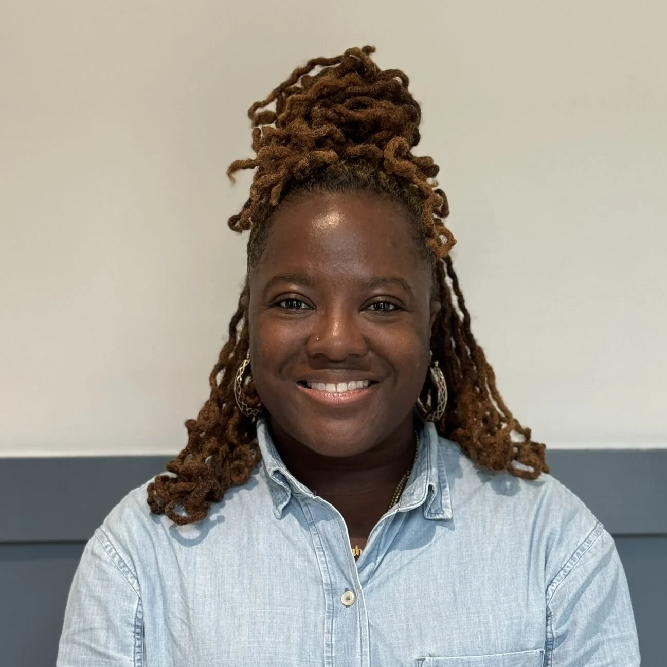 A woman with brown skin and blonde dreadlocks styled in a high bun, wearing a light blue denim shirt and gold hoop earrings, smiling at the camera.