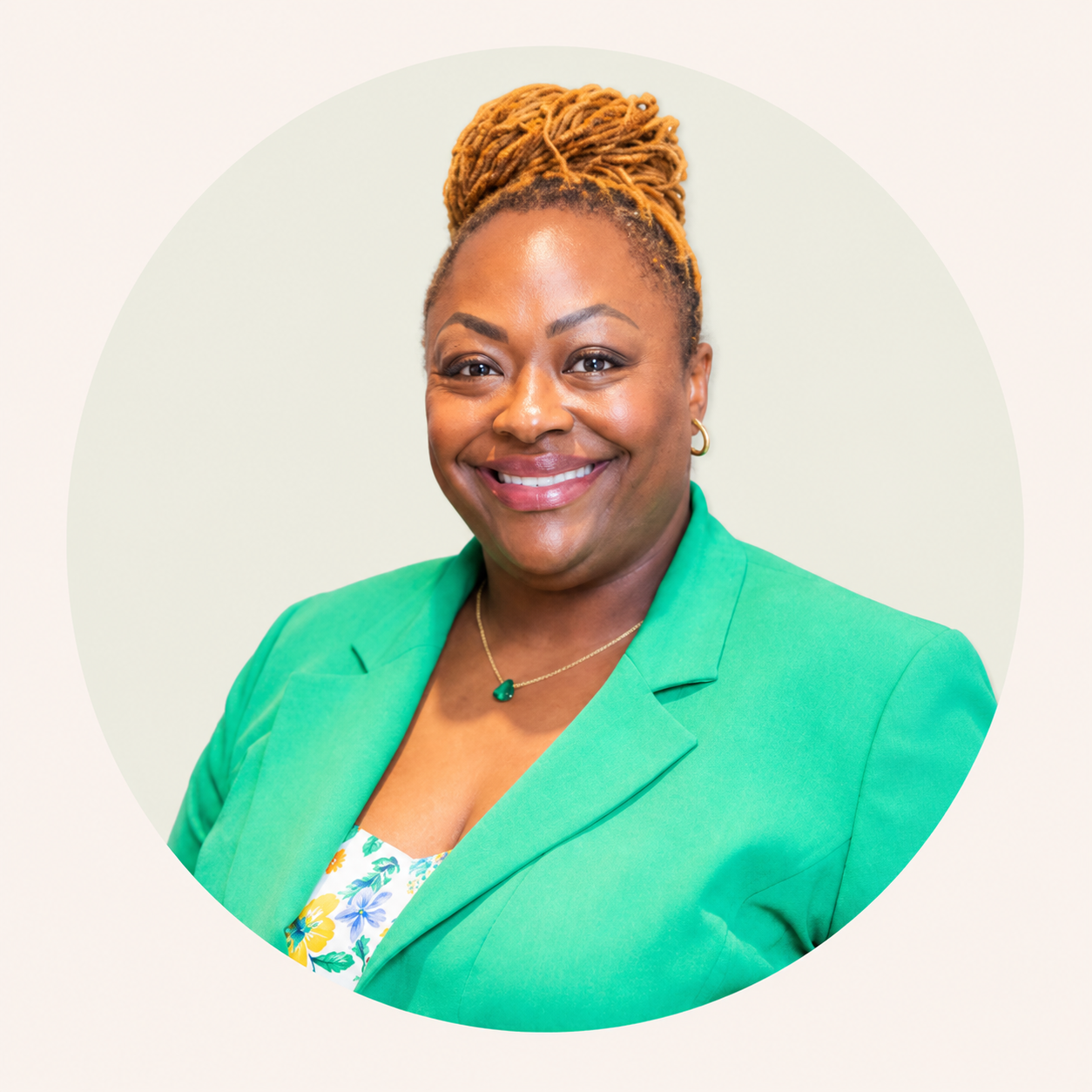 Portrait of an African American woman with dreadlocks in a bun, wearing a green blazer, a floral blouse, gold earrings, and a necklace with a green pendant, smiling at the camera.