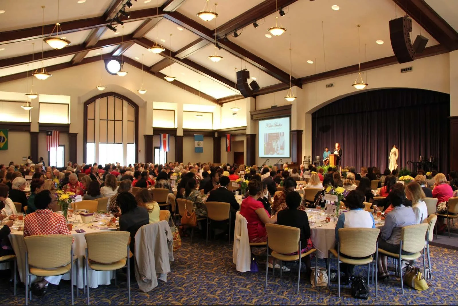 A large banquet hall filled with people seated at round tables during an event or conference. The room features high ceilings with wooden beams, large windows, and flags hanging on the wall. There is a stage at the front with a speaker, a large screen displaying a presentation, and a purple curtain backdrop.