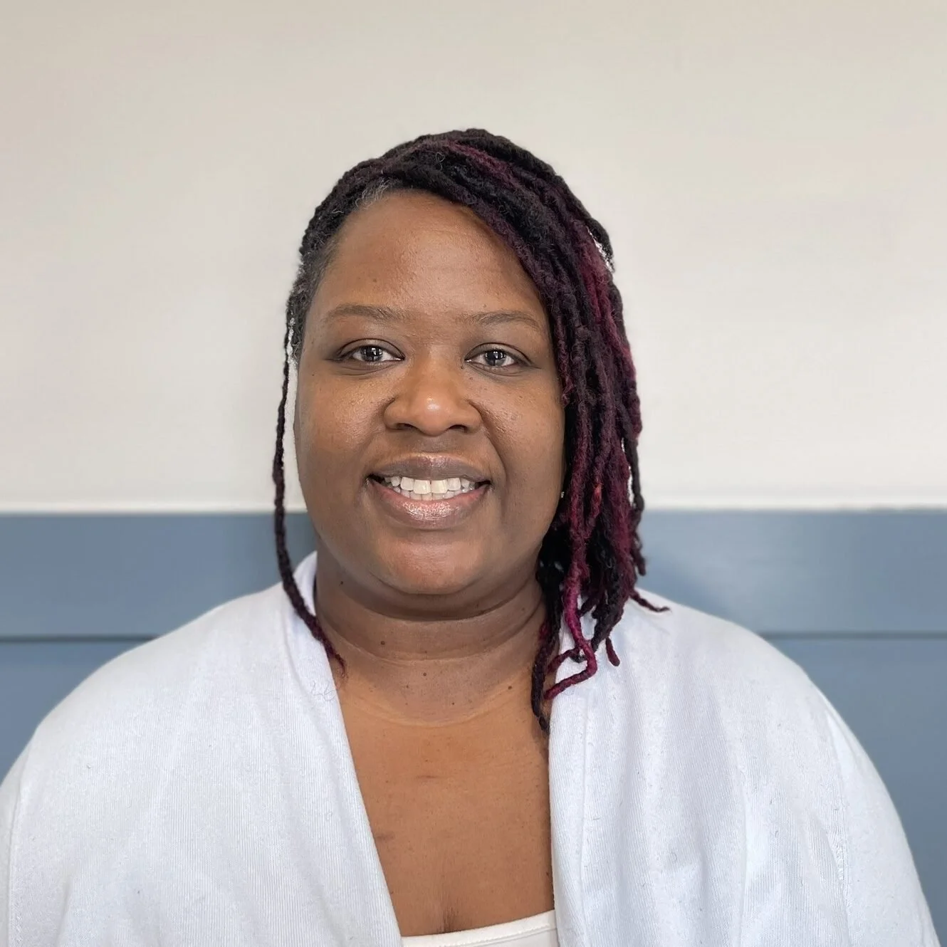 Portrait of a woman with short hair and purple dreadlocks, smiling, wearing a white shirt, against a plain background.