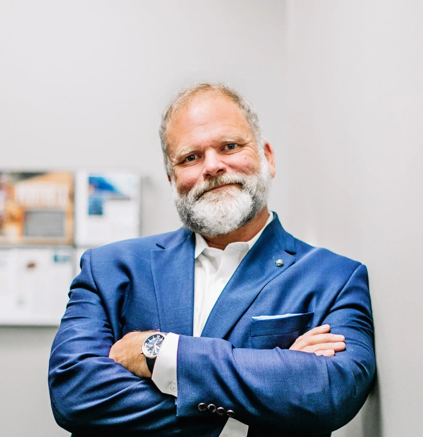 Man with gray hair and beard wearing a blue suit and white shirt, standing with arms crossed in front of a plain light-colored wall.