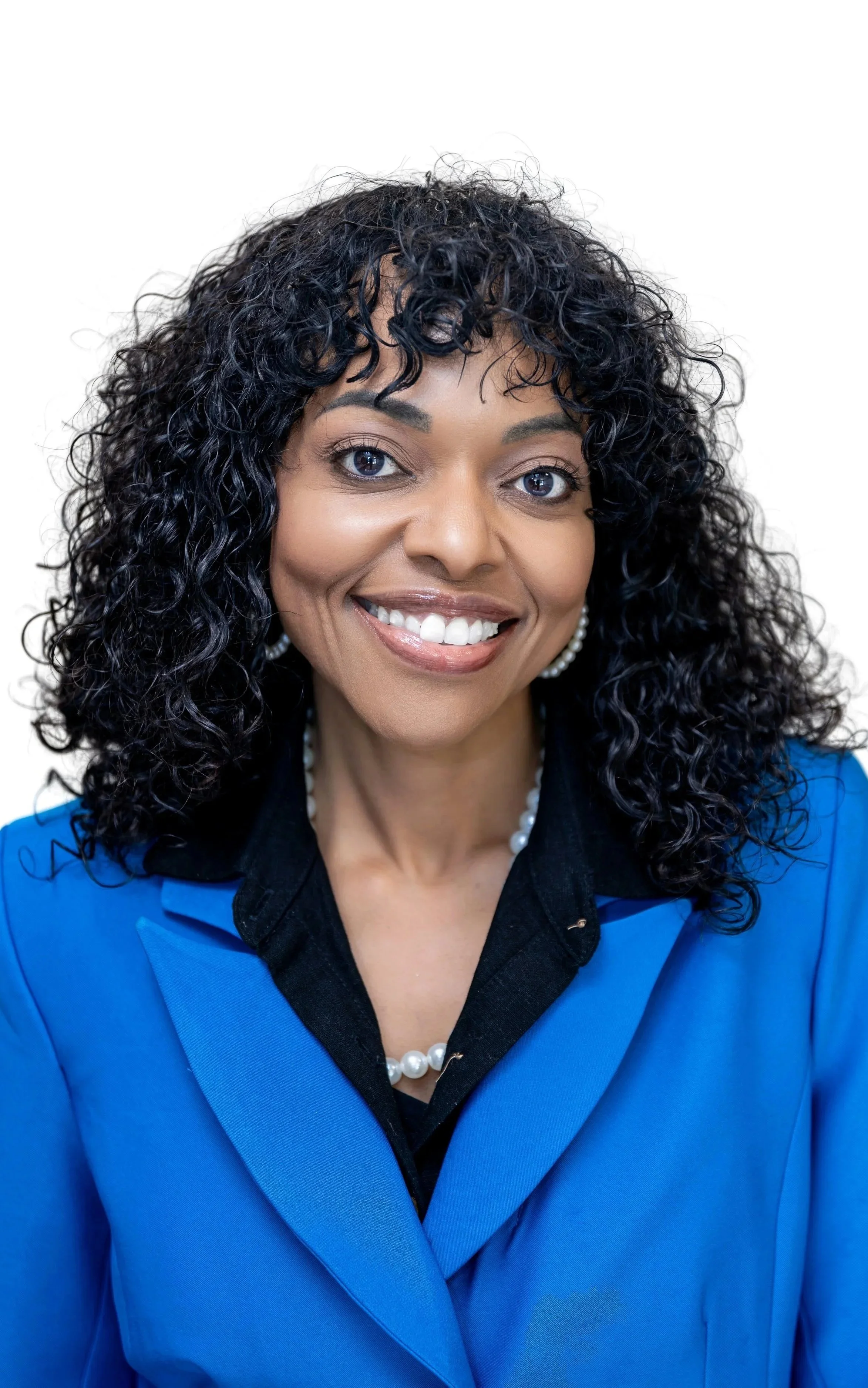Close-up of woman with curly black hair, smiling, wearing large gold hoop earrings and a colorful patterned shirt, against a plain light background.