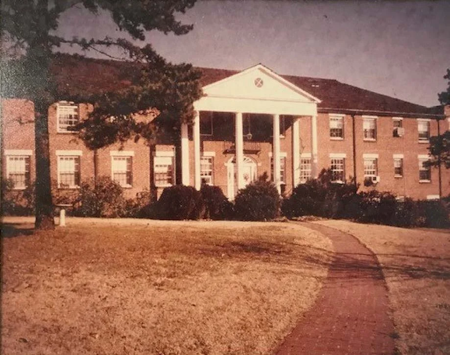 A large brick building with a white portico entrance and multiple windows, surrounded by trees and a curved brick pathway leading to the entrance, on a sunny day.