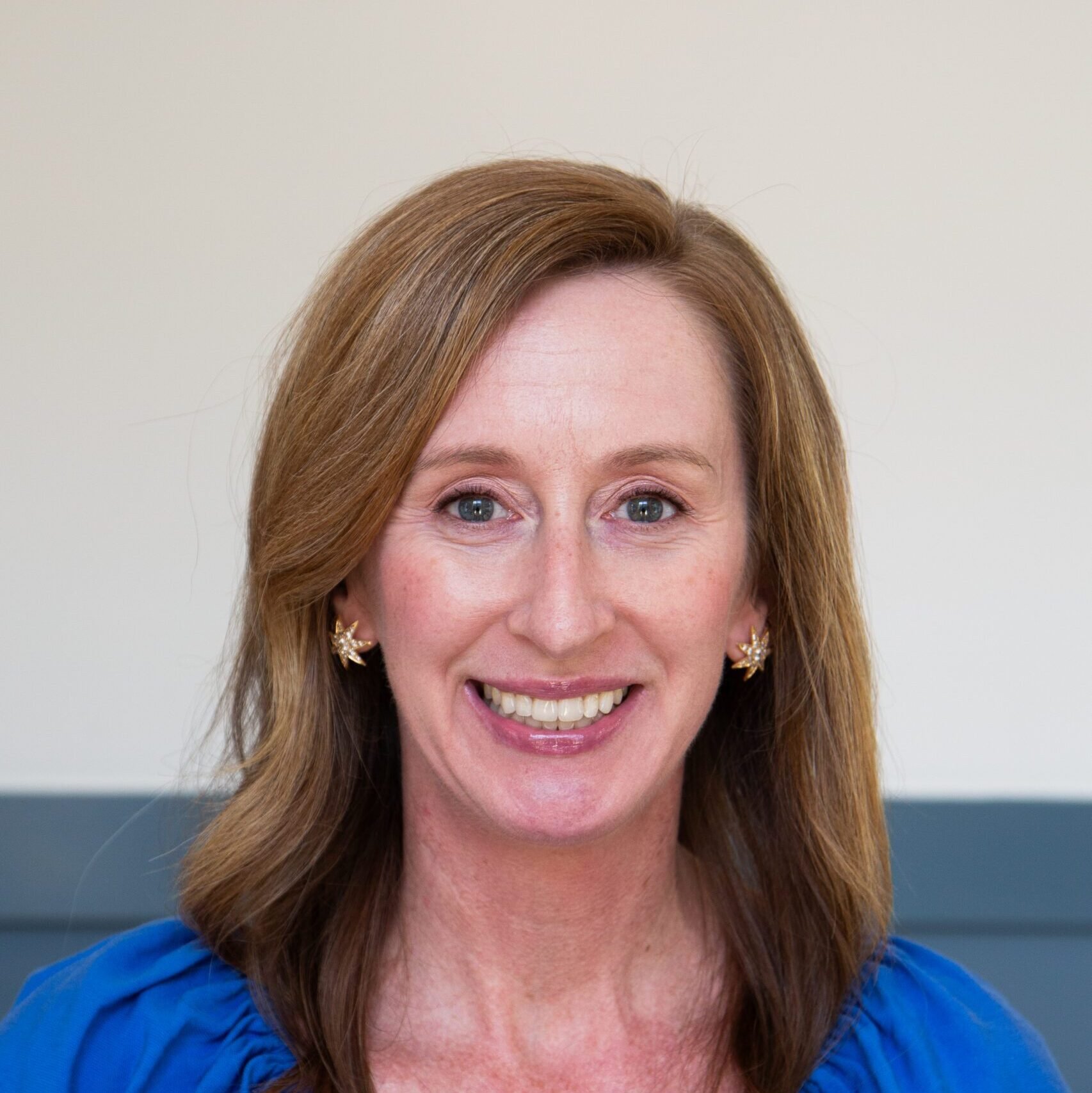 Close-up of a woman with light skin, brown hair, blue eyes, smiling, wearing gold floral earrings, blue top, against a plain light background.