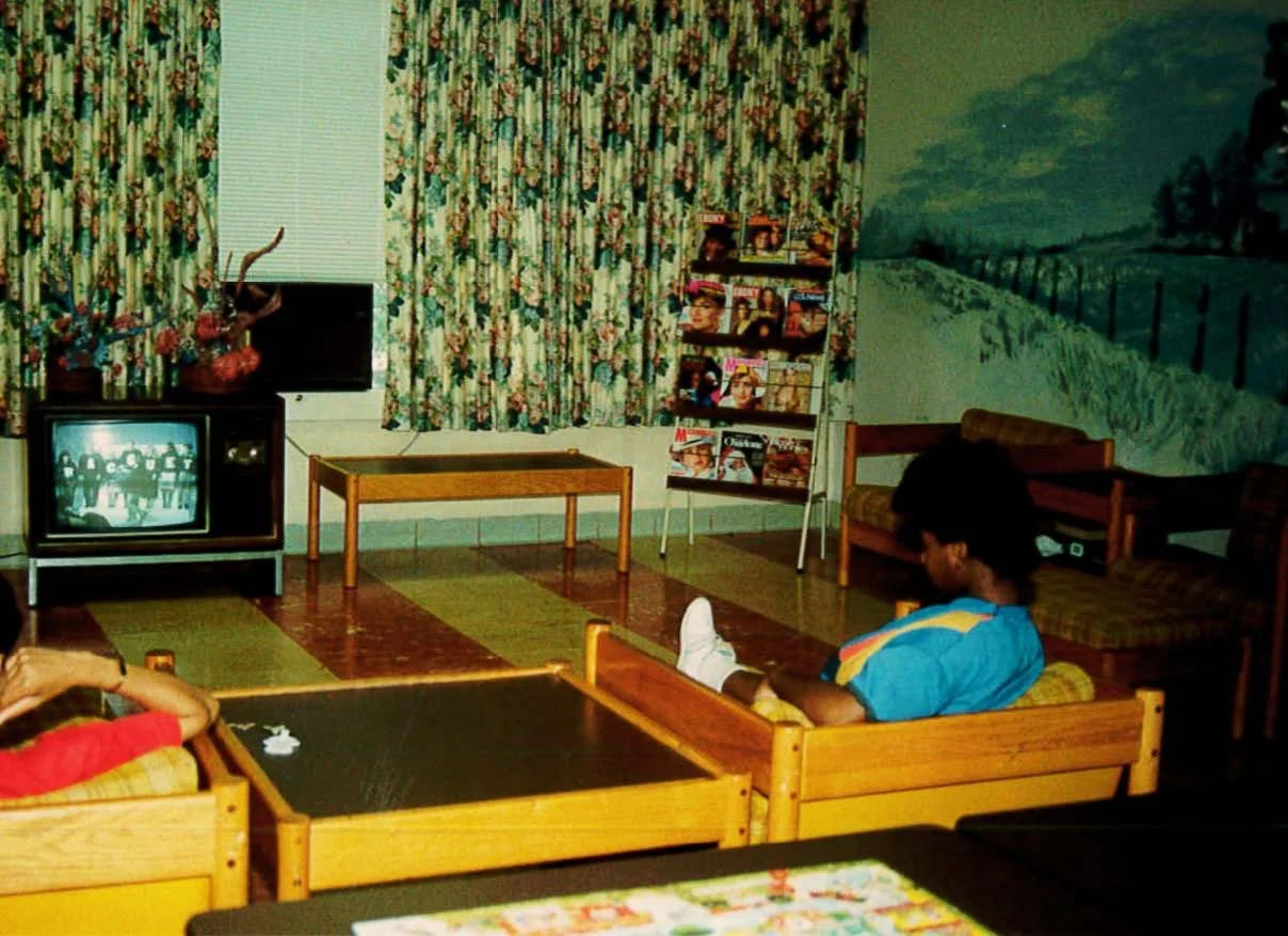 A living room with vintage decor, two children watching TV, floral curtains, magazine rack, and a mural of a landscape on the wall.
