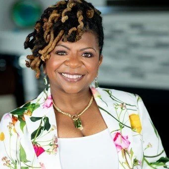 A woman smiling with short curly hair and wearing a floral blazer and gold jewelry, standing indoors.