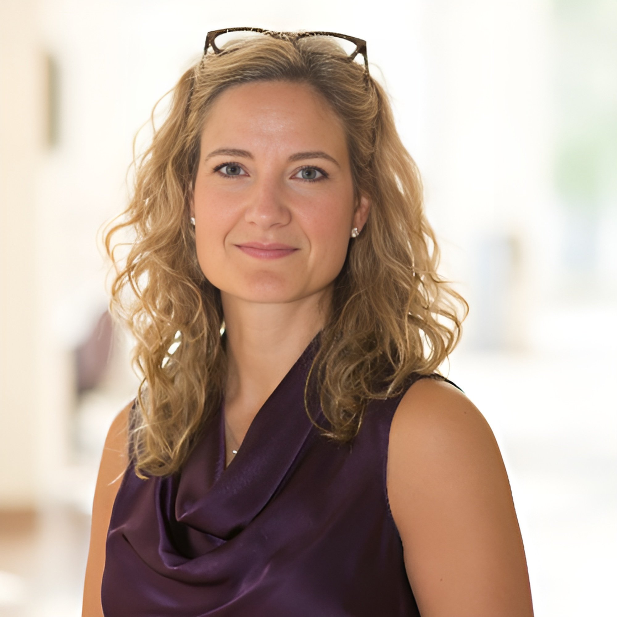 A woman with curly blonde hair, wearing a dark purple sleeveless top, and glasses resting on her head, smiling.