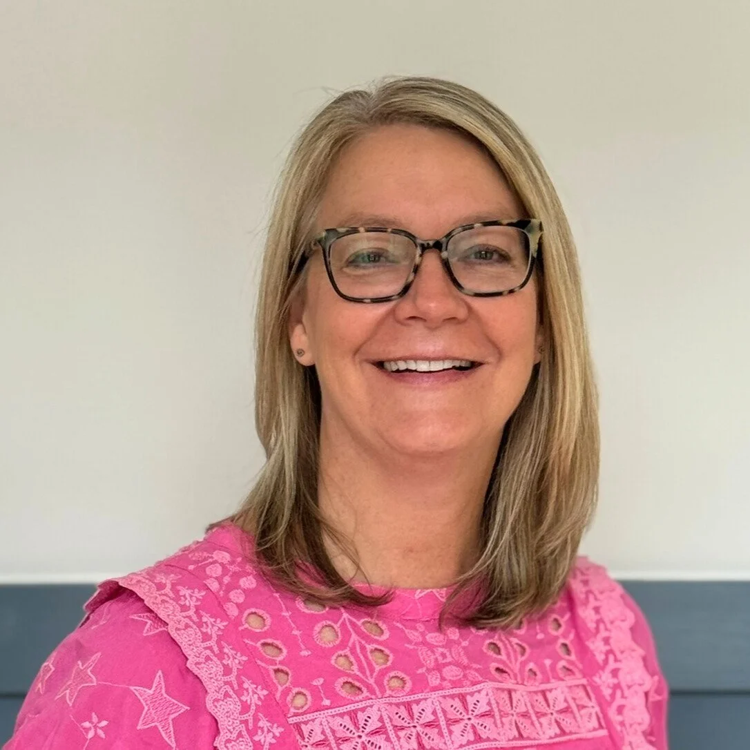 Smiling woman with blonde hair, glasses, and a pink embroidered top standing against a plain light-colored background.