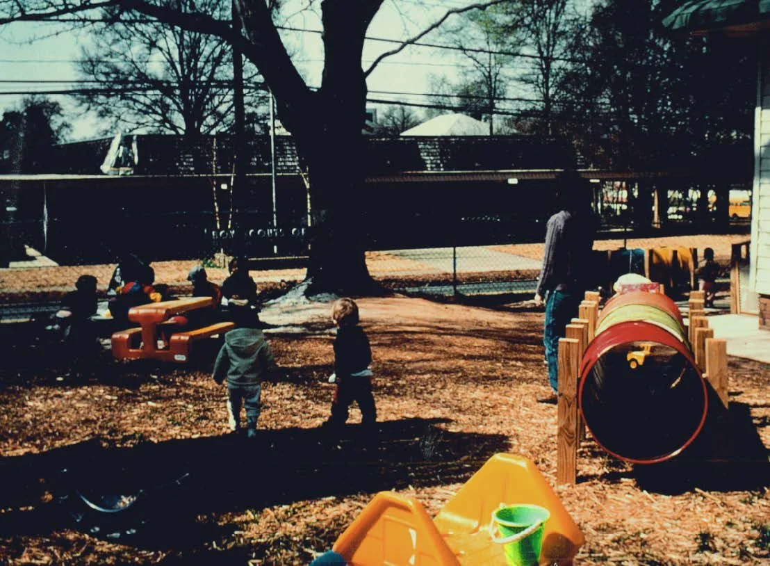 Children playing on playground equipment near a large tree in a park, with a person watching and a train in the background.