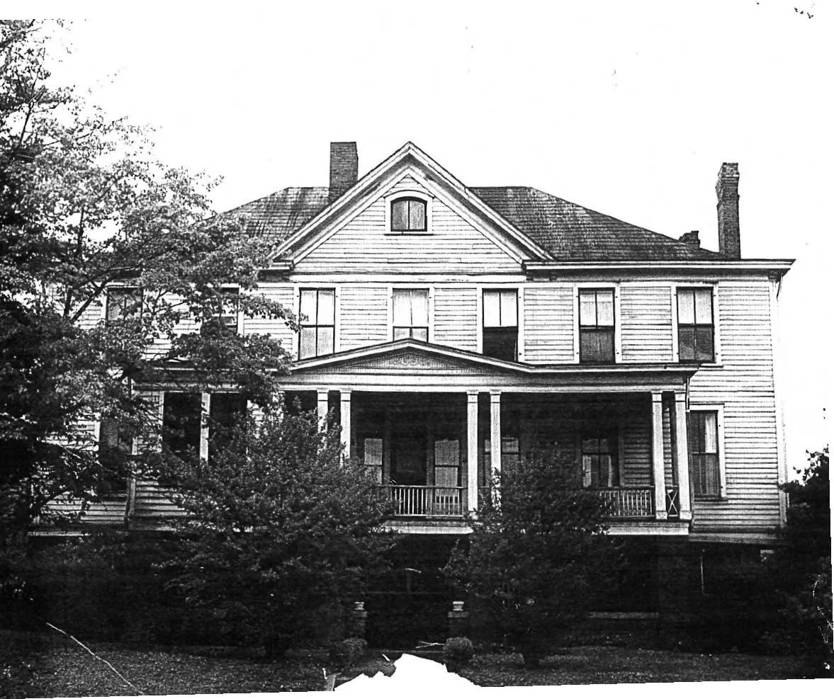 Black and white photo of a large, old, wooden house with a gabled roof, multiple chimneys, and a front porch with columns, surrounded by trees.