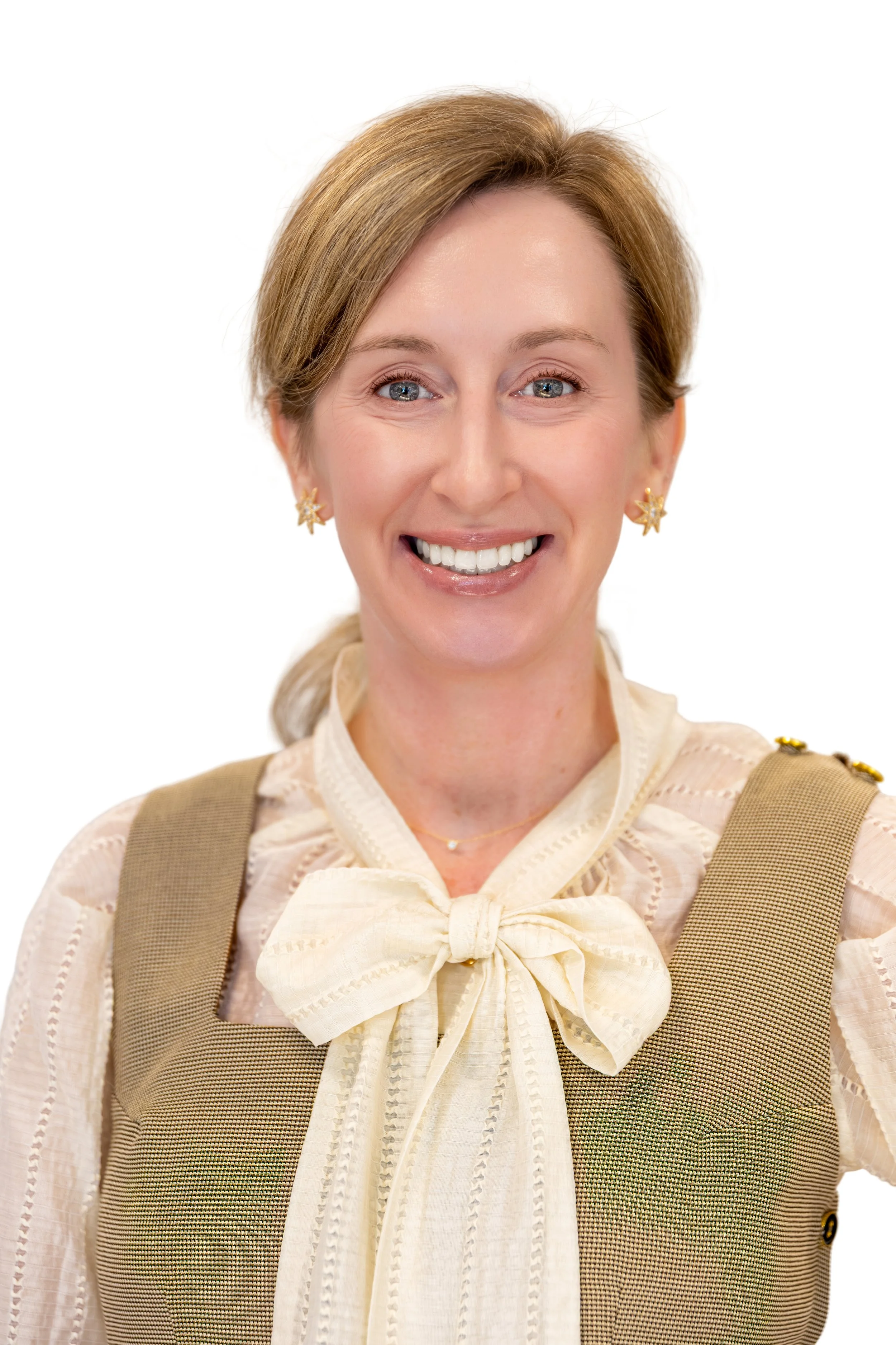 Close-up of a woman with light skin, brown hair, blue eyes, smiling, wearing gold floral earrings, blue top, against a plain light background.