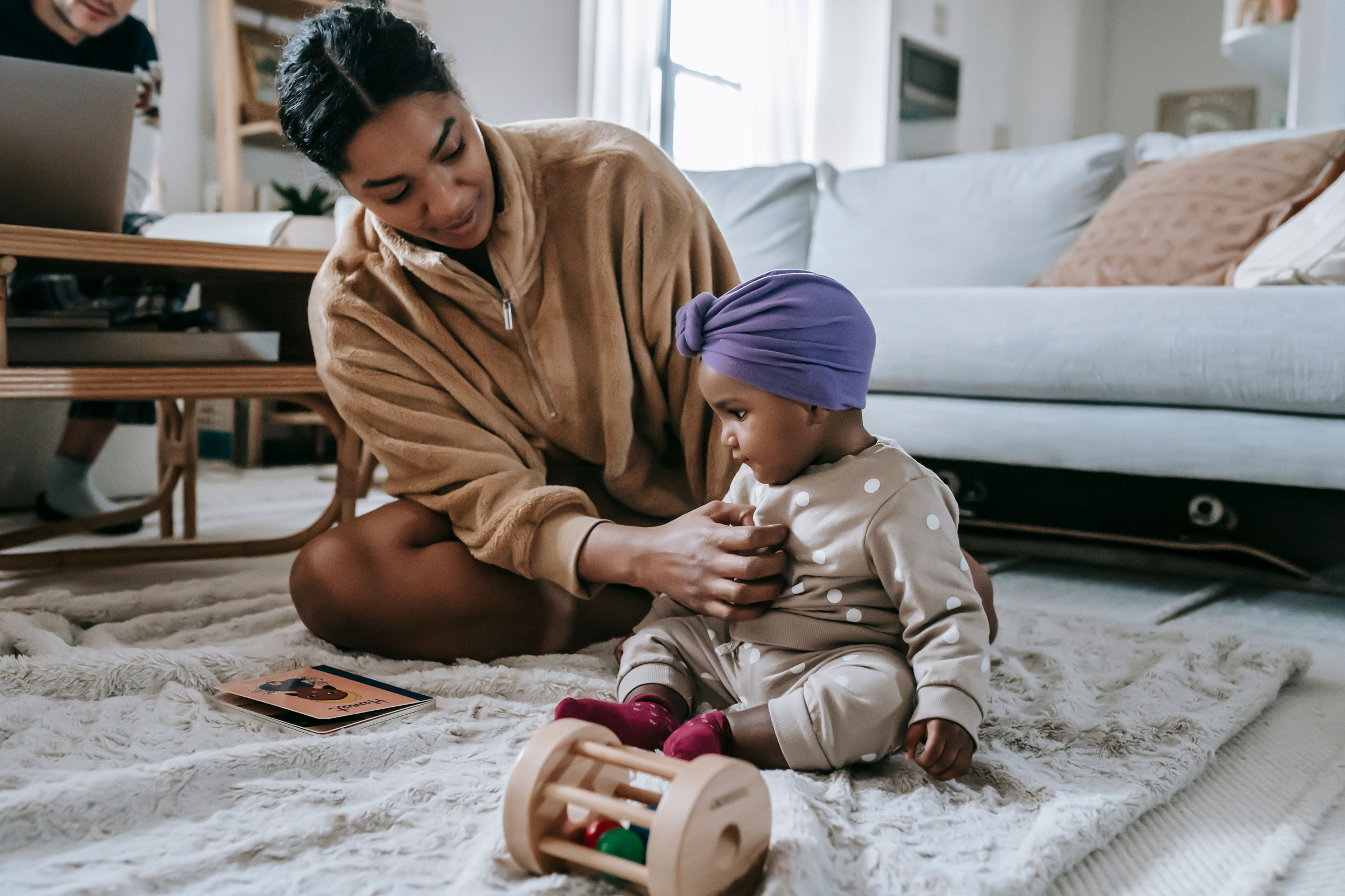 A woman and a young girl sitting on a soft, cream-colored shaggy rug in a cozy living room. The woman is leaning forward, wearing a tan fleece jacket, and the girl is sitting cross-legged, wearing beige pajamas with white polka dots and a purple head wrap. There are children's toys near them and a magazine on the rug. In the background, there is a couch with beige and white pillows, a coffee table, and a person in dark clothing working on a laptop.