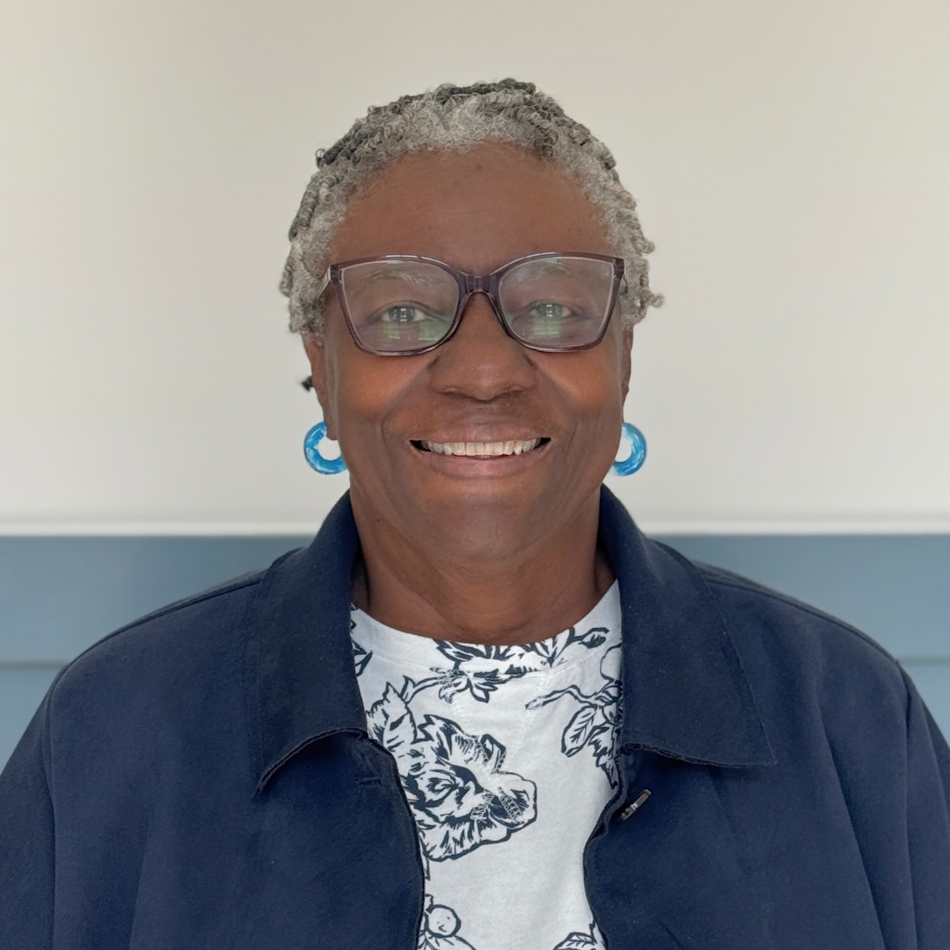 Smiling woman with gray twisted hair, wearing glasses, large blue earrings, a dark jacket, and a white shirt with black leaf print, standing against a plain light background.