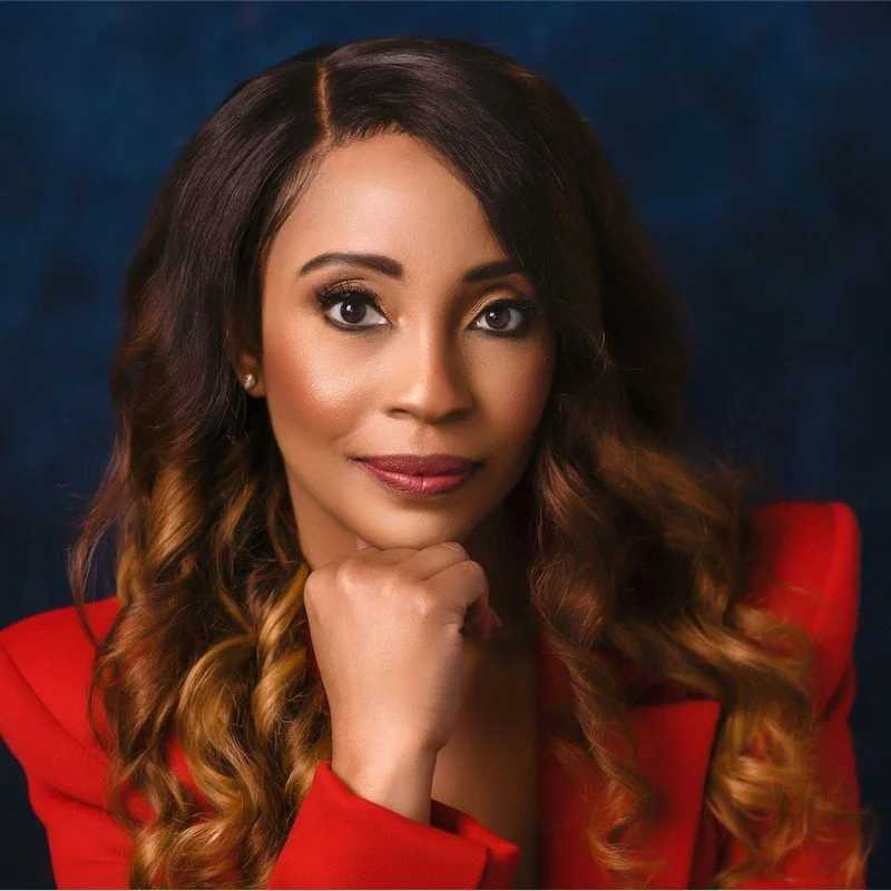 A woman with long, wavy hair wearing a red blazer, posing with her hand under her chin against a dark blue background.