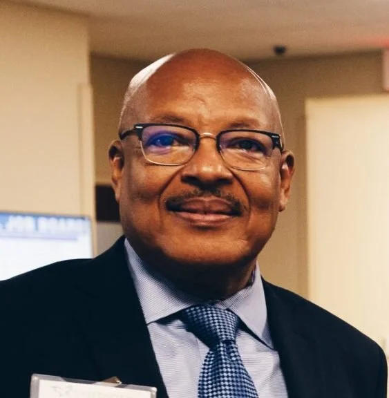 A smiling man in formal attire, wearing glasses, in an indoor setting.