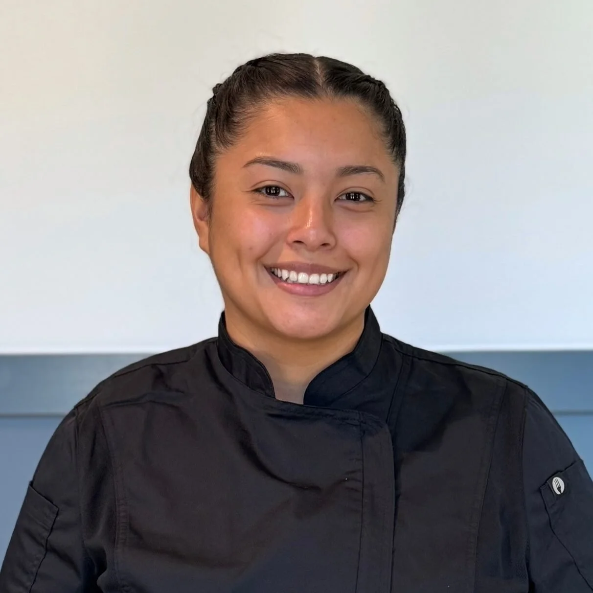A woman in a black chef's coat smiling in front of a plain light-colored wall.