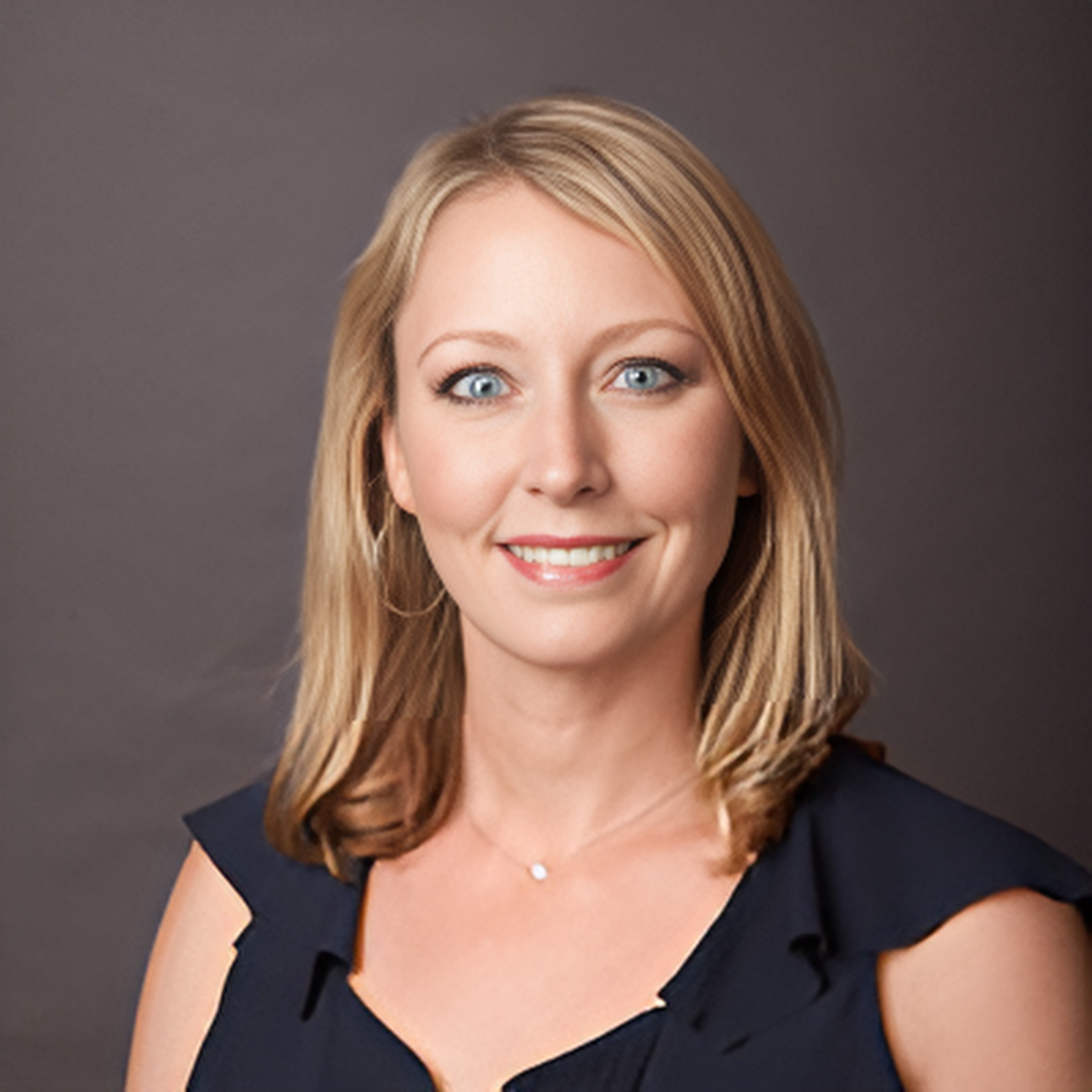 Professional headshot of a woman with blonde hair, blue eyes, smiling, wearing a dark dress with ruffle sleeves, against a dark grey background.