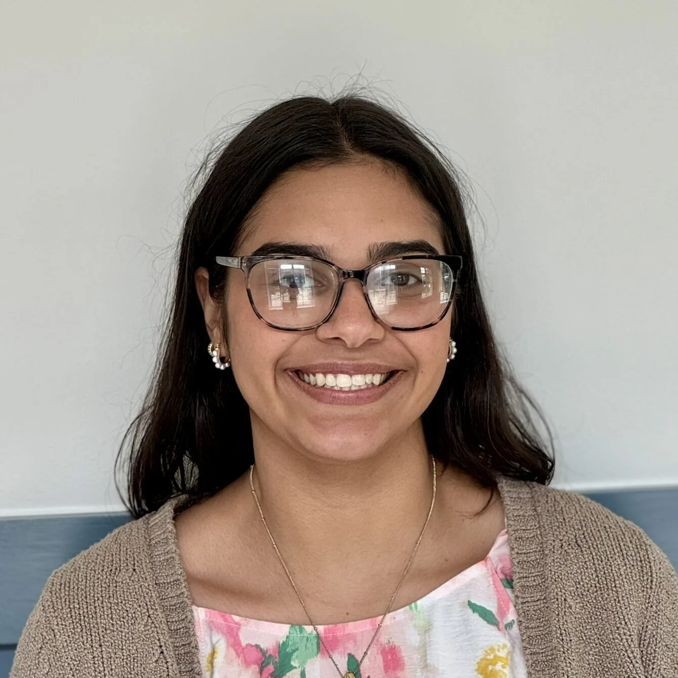 A woman with dark hair, glasses, and jewelry smiling in front of a light-colored wall.