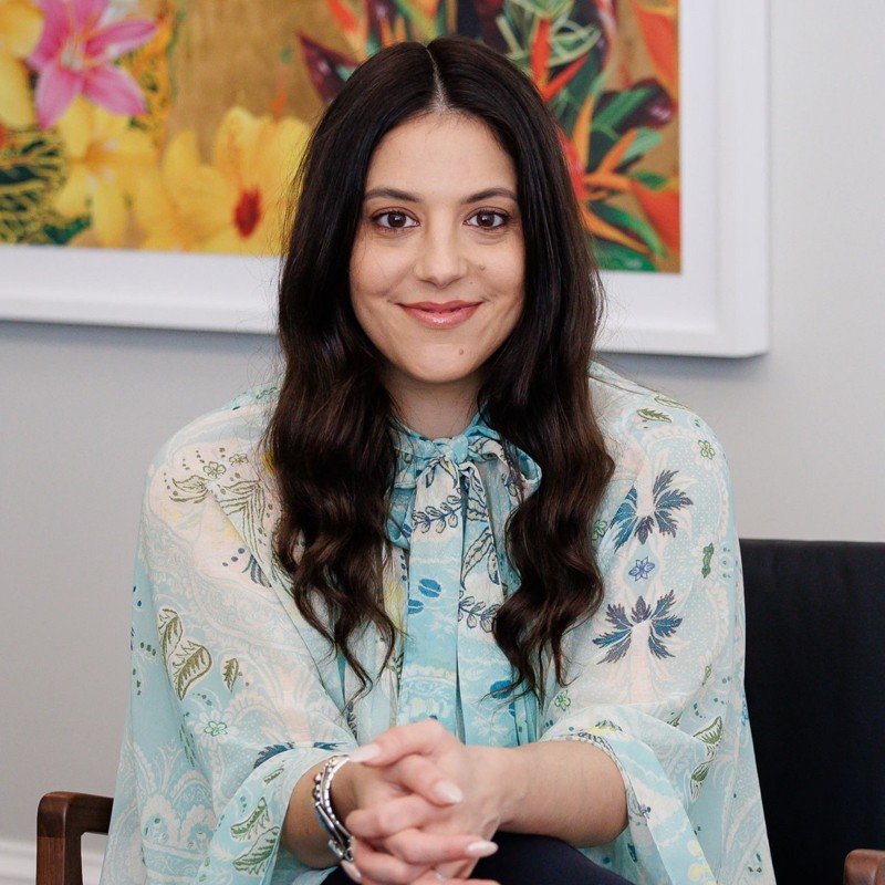 A woman with long dark hair and light skin is sitting at a table, smiling, with a colorful floral art piece in the background.