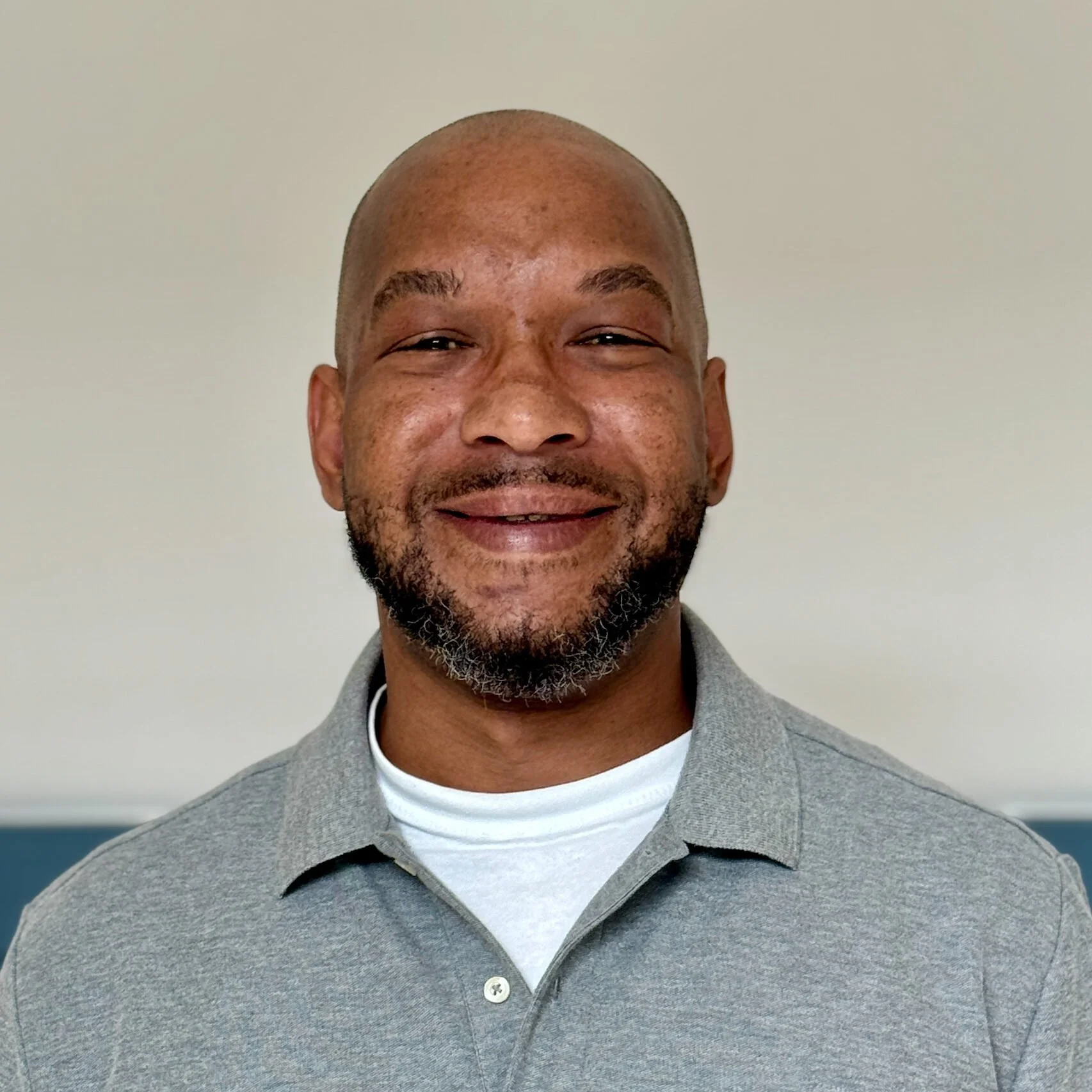 Close-up of a smiling man with a shaved head and a beard, wearing a grey collared shirt and a white undershirt, standing against a plain light-colored wall.