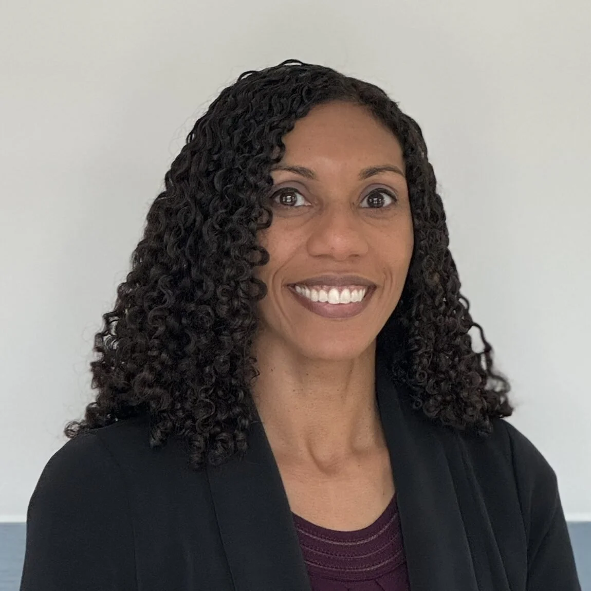 A woman with curly black hair smiling, wearing a black blazer and maroon top, against a plain white background.