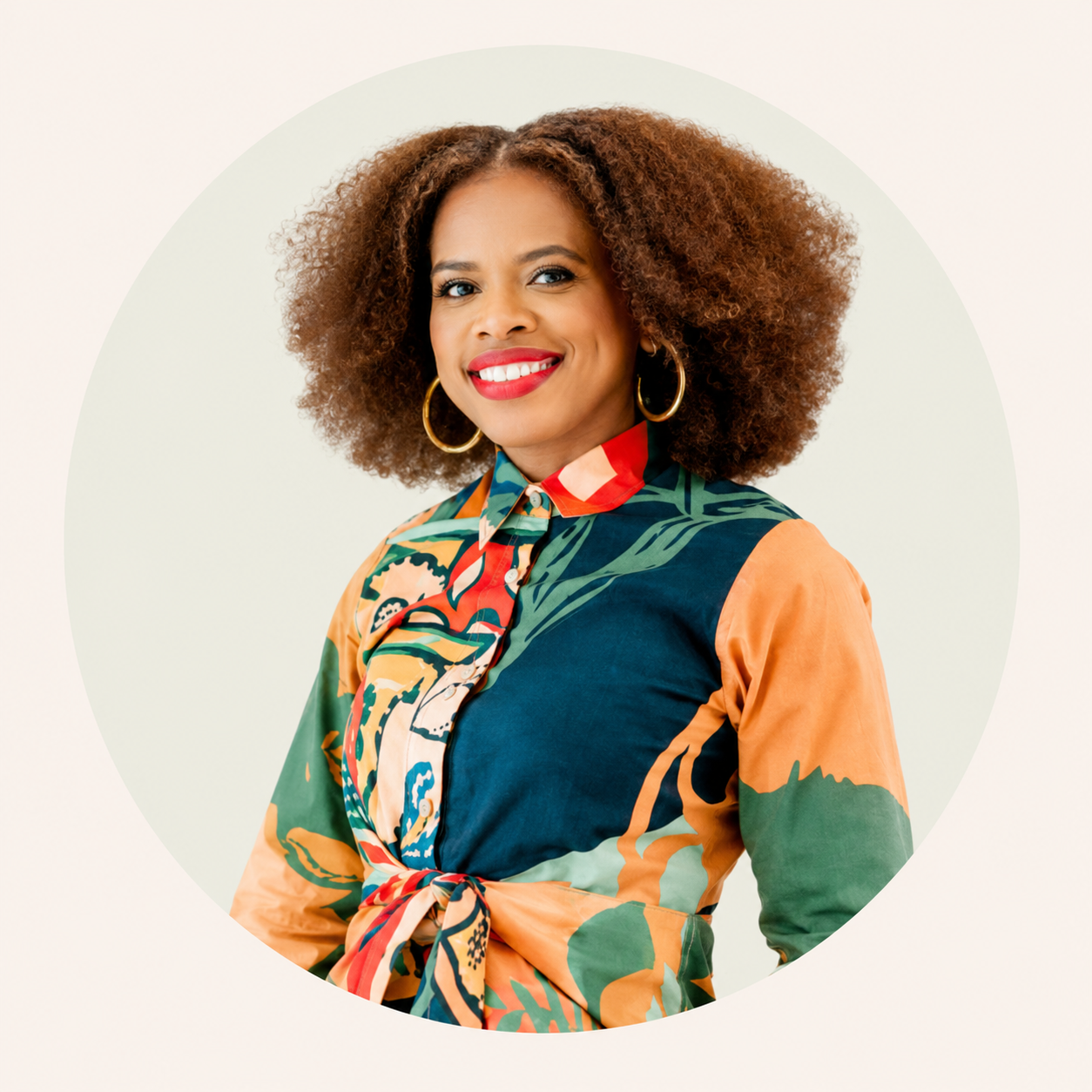 Portrait of a smiling woman with curly hair, wearing gold hoop earrings and a colorful patterned blouse tied at the waist, against a light background with a circular design.