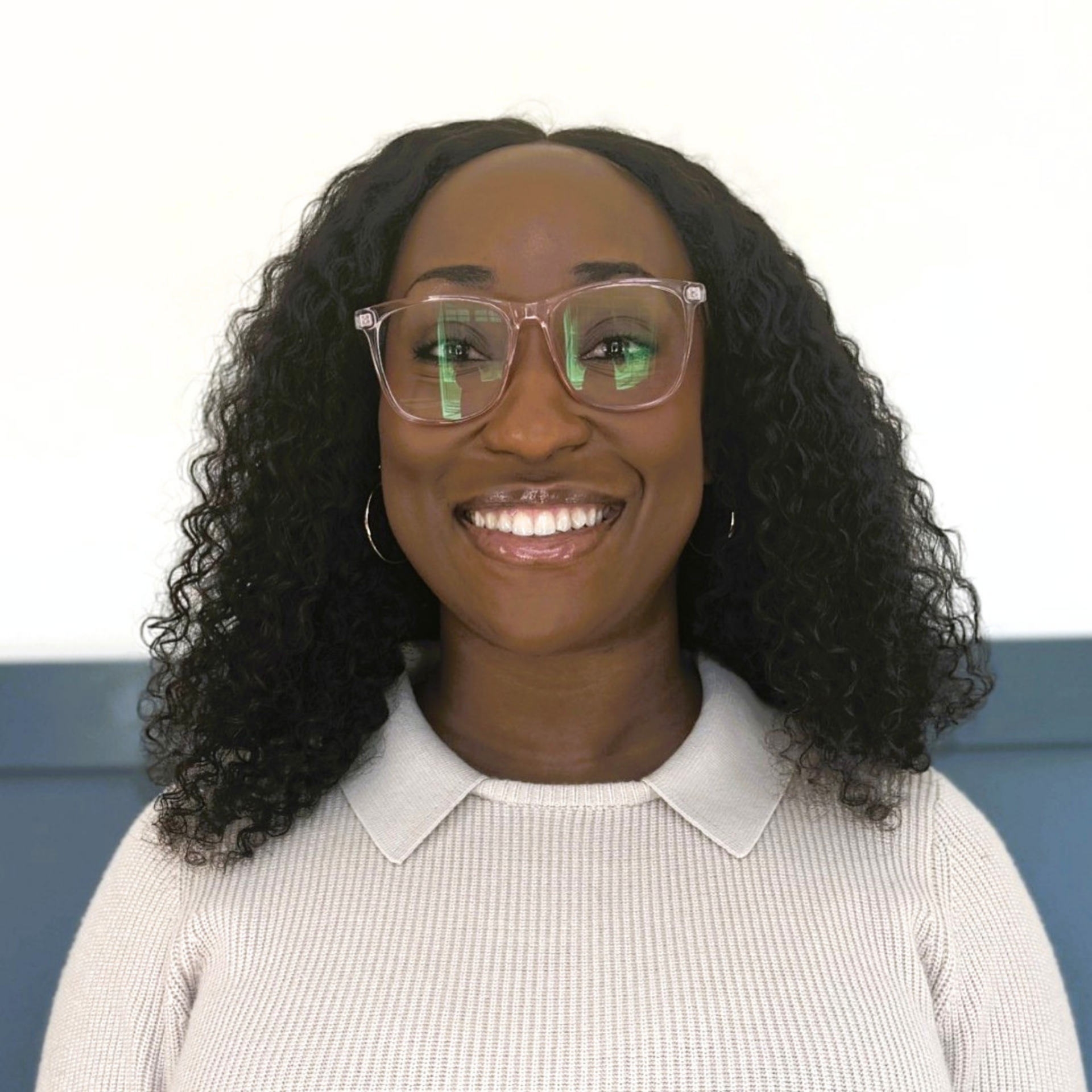 Close-up of a smiling woman with dark, curly hair wearing clear glasses and a light-colored sweater against a plain background.