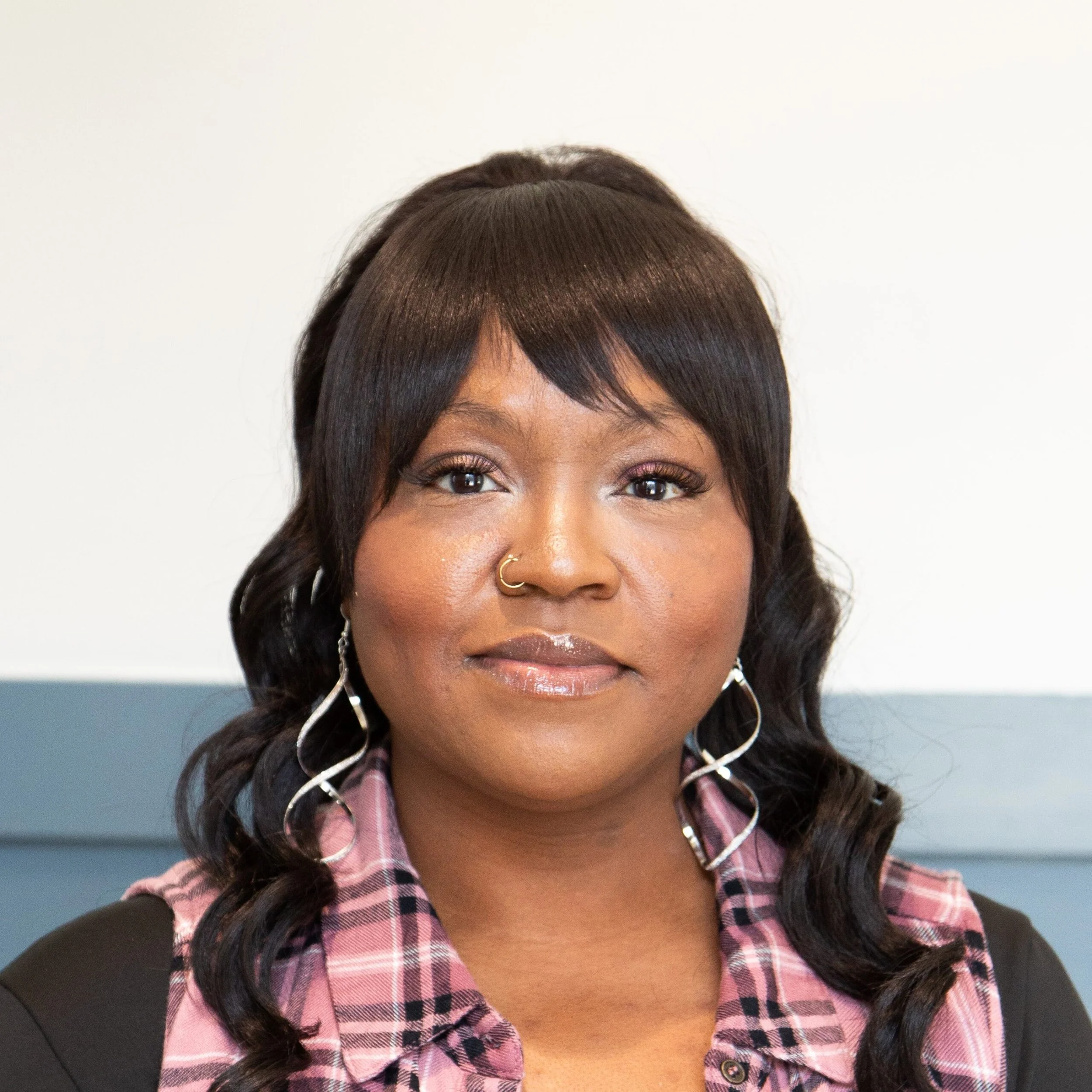 Close-up of a woman's face showing her eyes, nose, lips, and earrings, with her hair styled in loose waves and a side braid, wearing a pink and black plaid shirt.