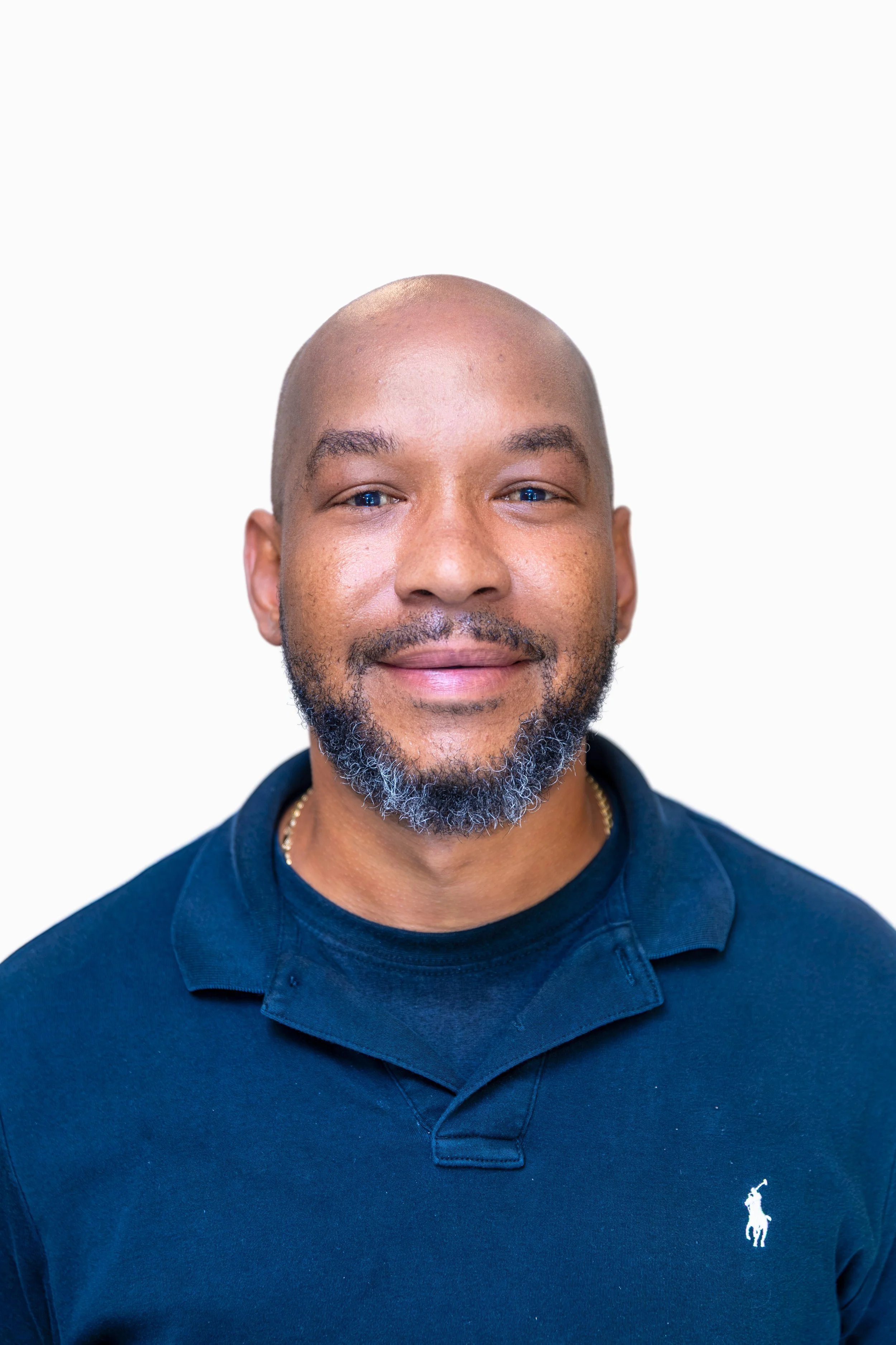 Close-up of a smiling man with a shaved head and a beard, wearing a grey collared shirt and a white undershirt, standing against a plain light-colored wall.
